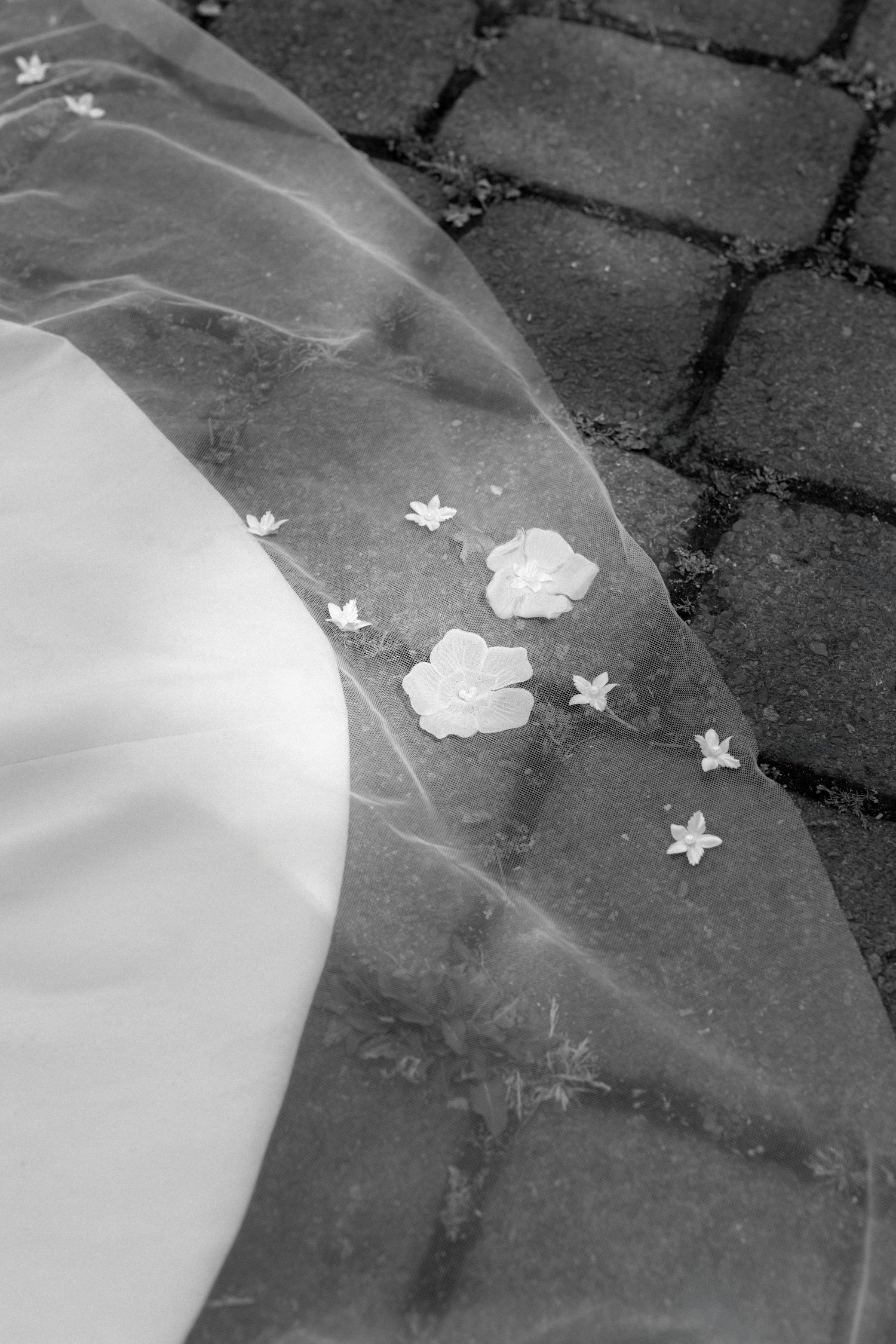 Close-up black and white photo of a wedding dress train with delicate floral details on a cobblestone path.