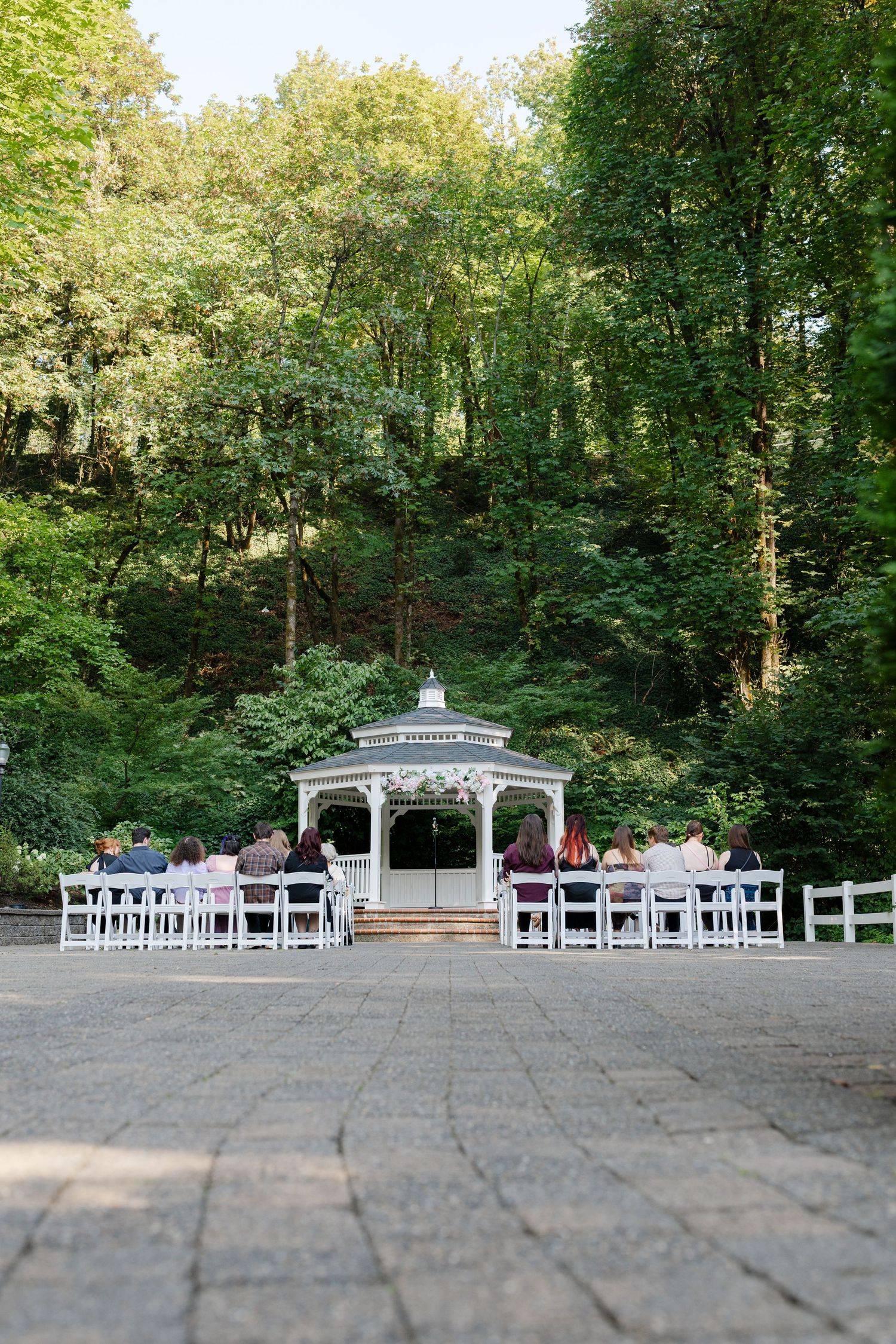 Outdoor wedding ceremony with guests seated in white chairs facing decorated gazebo in wooded setting.
