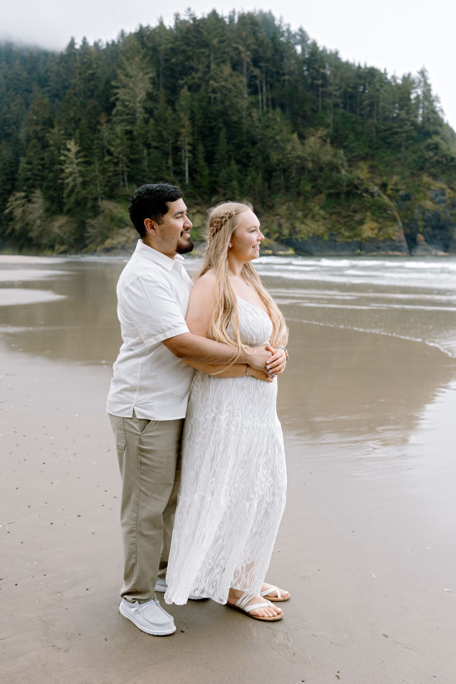 Couple embracing on a sandy beach with lush green forest backdrop, woman in white lace dress and man in white shirt.