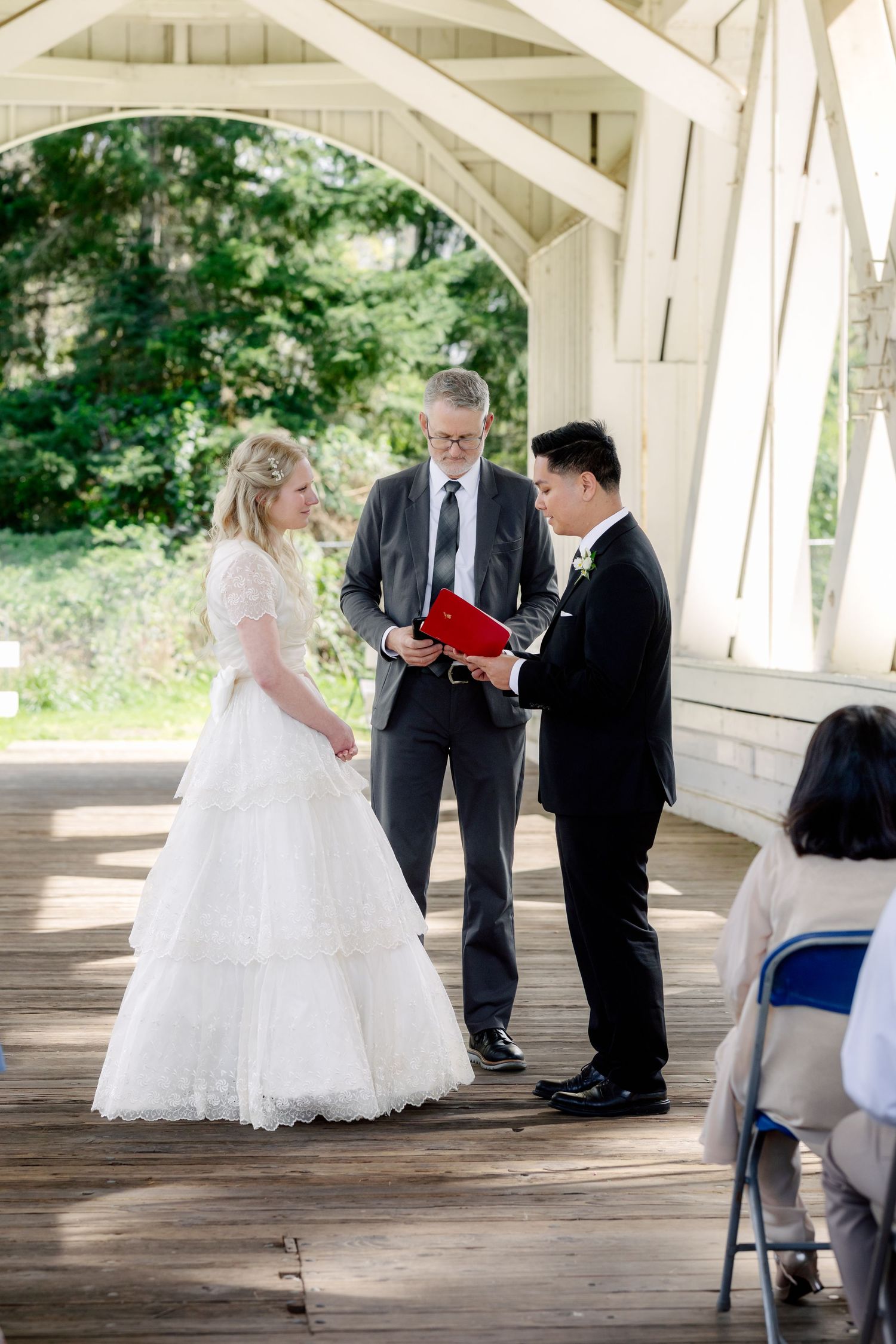 Bride and groom exchange vows under a white covered bridge pavilion during an outdoor wedding ceremony.