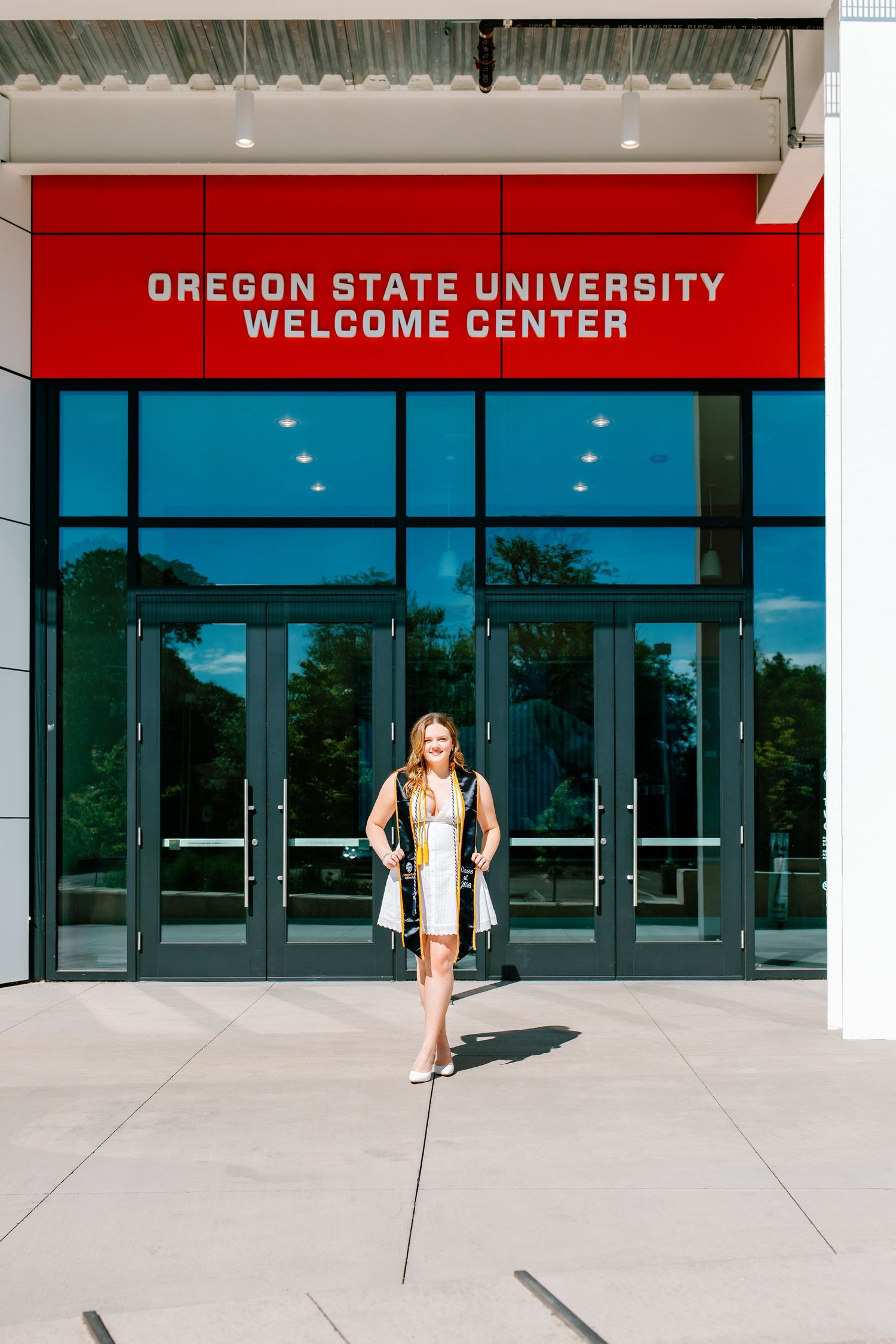 Female graduate in white dress and black graduation stole stands before Oregon State University Welcome Center.