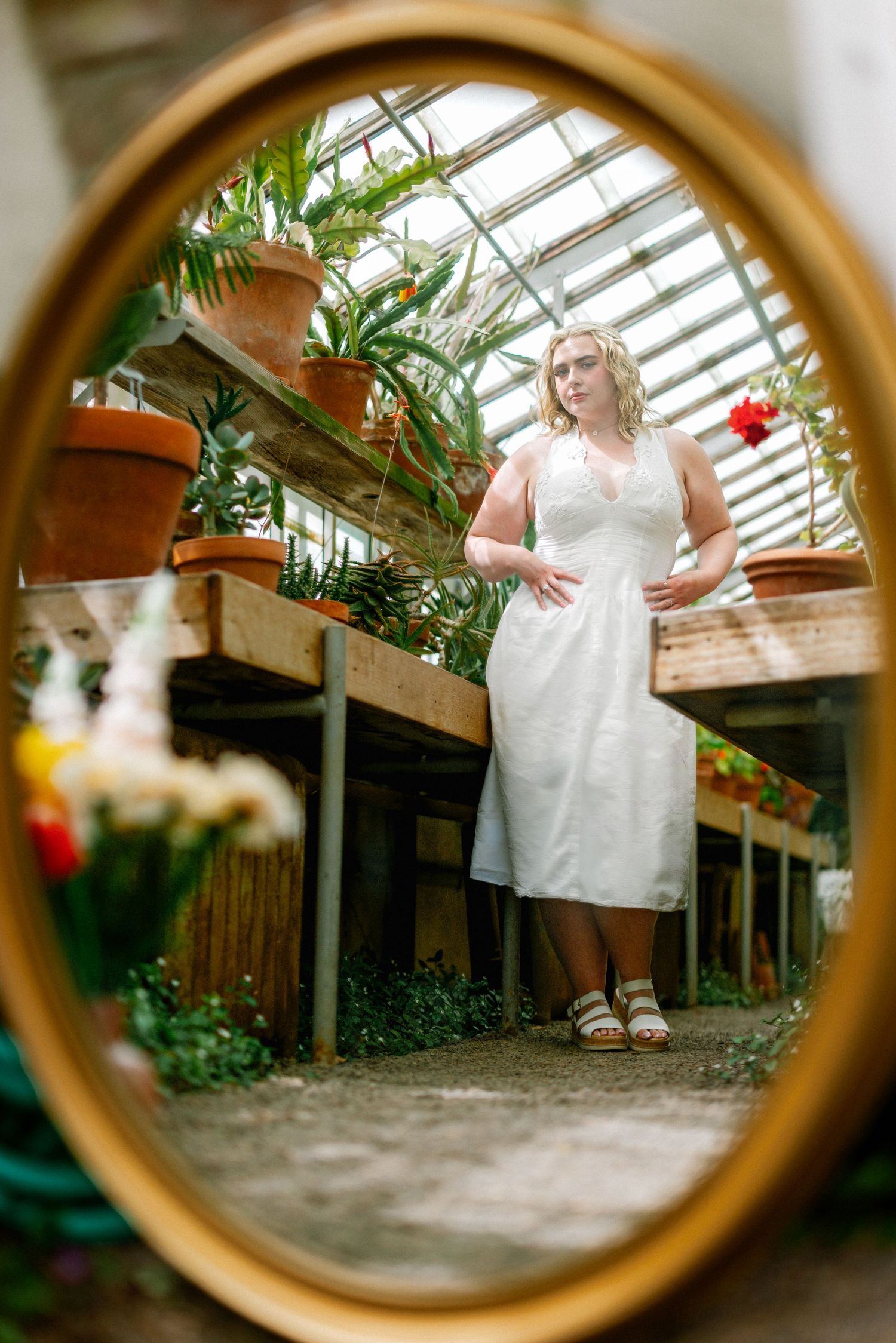 Woman in white dress reflected in oval mirror inside a lush greenhouse with potted plants and glass ceiling.