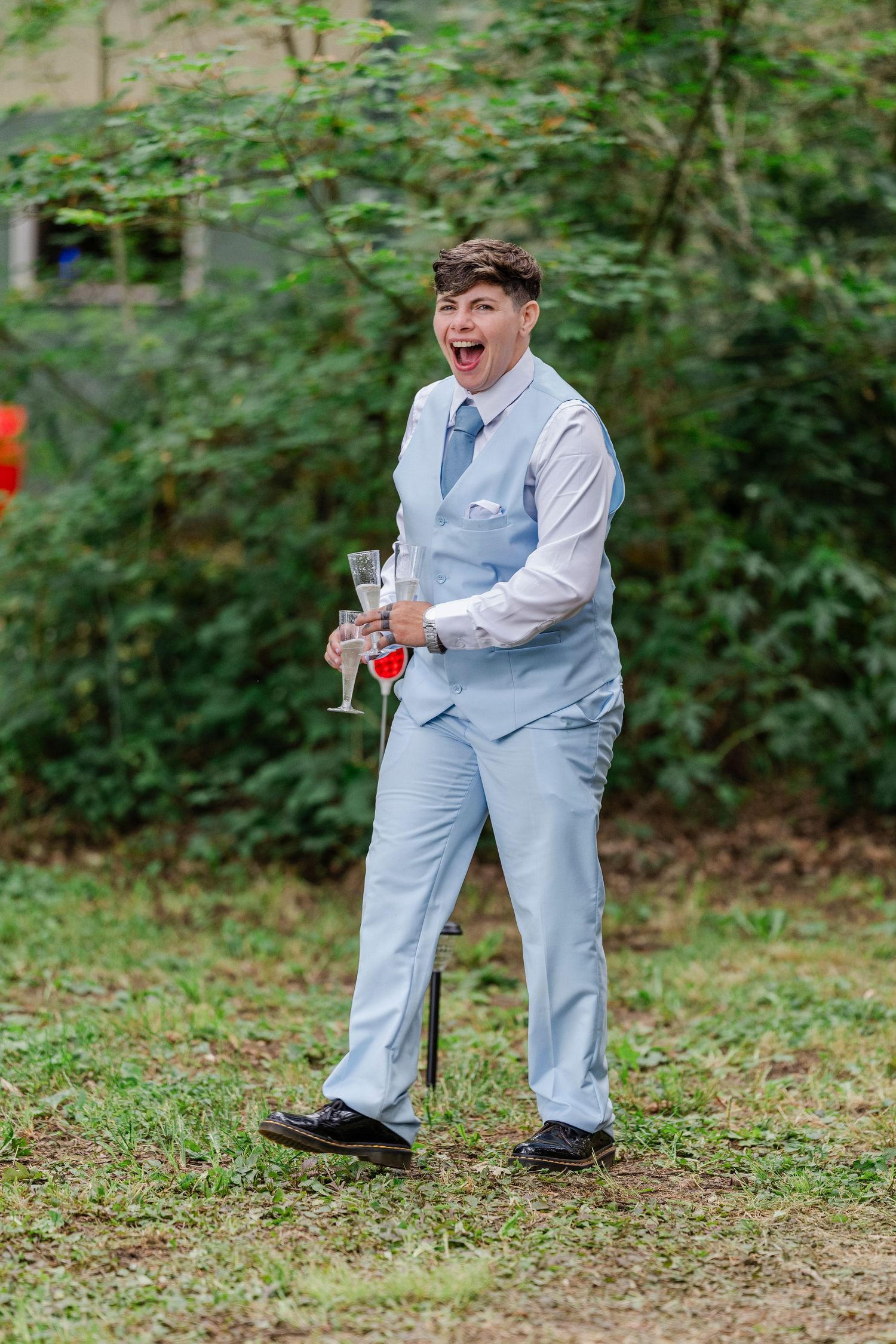 Someone in a light blue suit vest and tie laughs joyfully while holding a champagne glass at an outdoor event.
