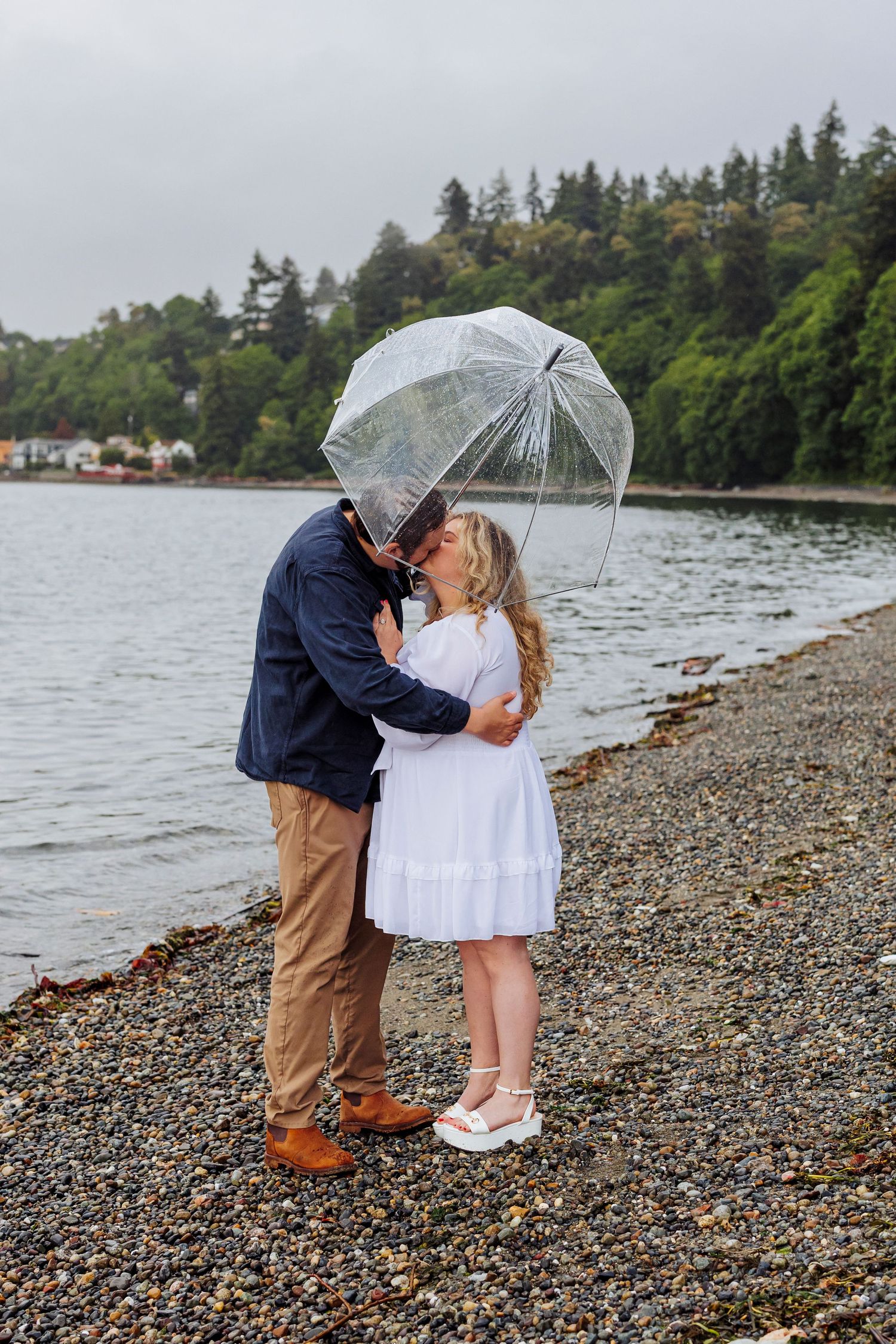 A couple shares a romantic kiss under a clear umbrella on a rocky beach shore with evergreen trees in the background.