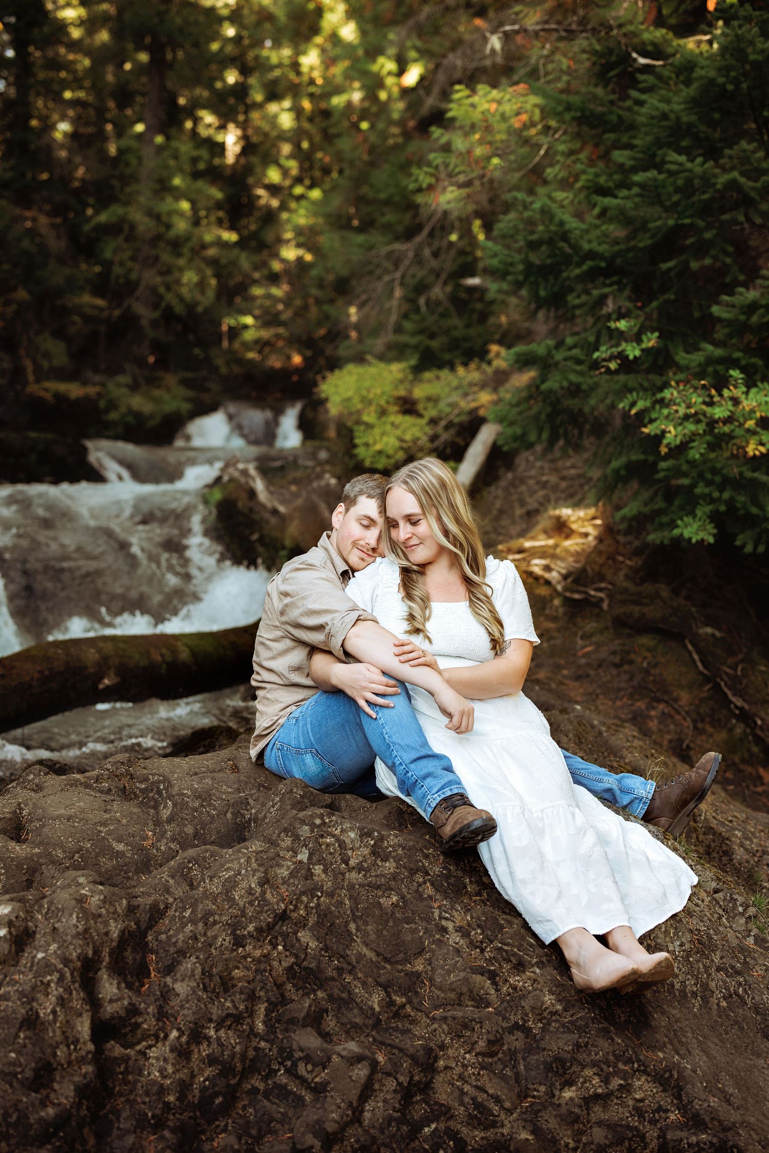 Two people lounging on large rocks near waterfall in forest setting during romantic photoshoot.