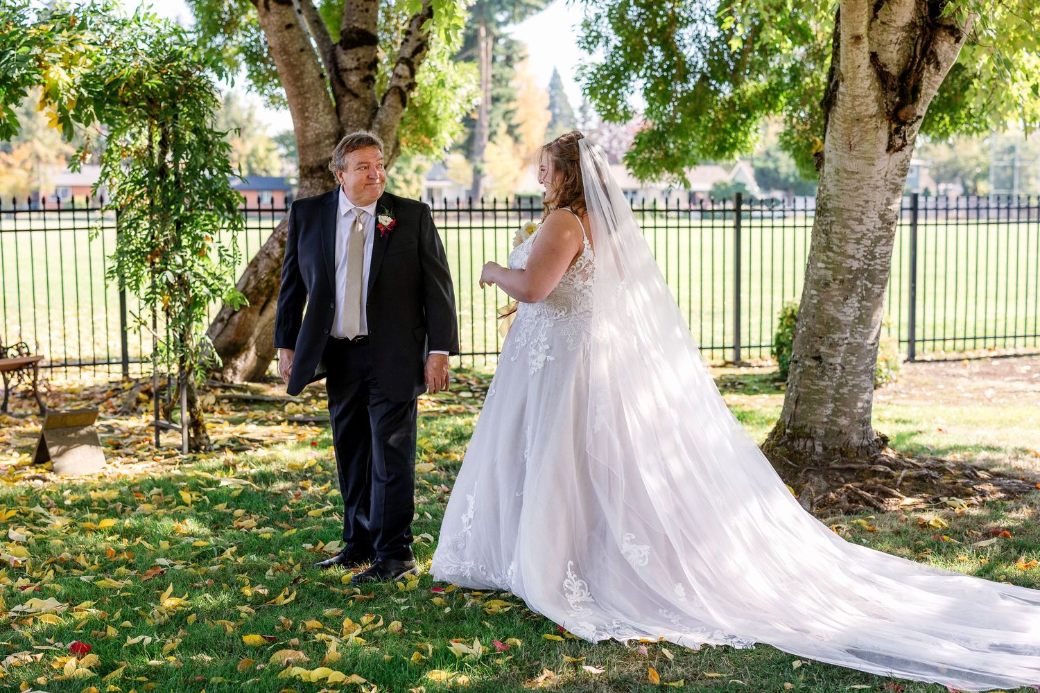 A couple in wedding attire walking together under trees in a park with a black iron fence in the background.