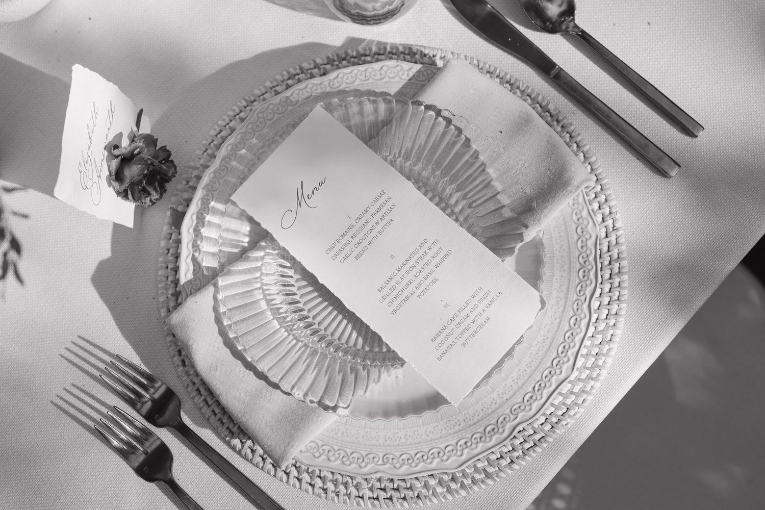 Black and white photo of elegant place settings with silver cutlery and ornate plates on a formal dining table.