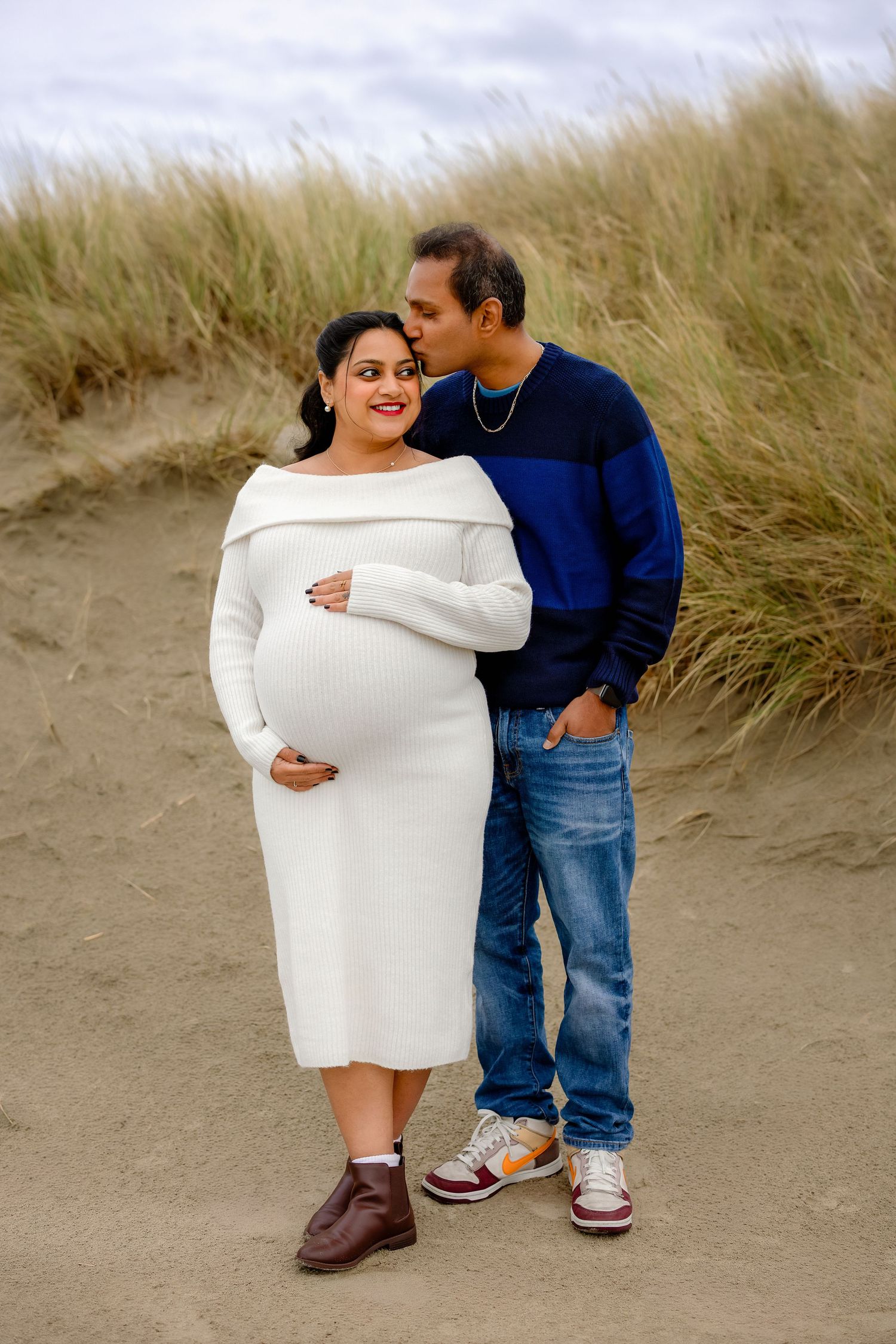 A couple shares an intimate moment on a sandy beach with coastal grass in the background.