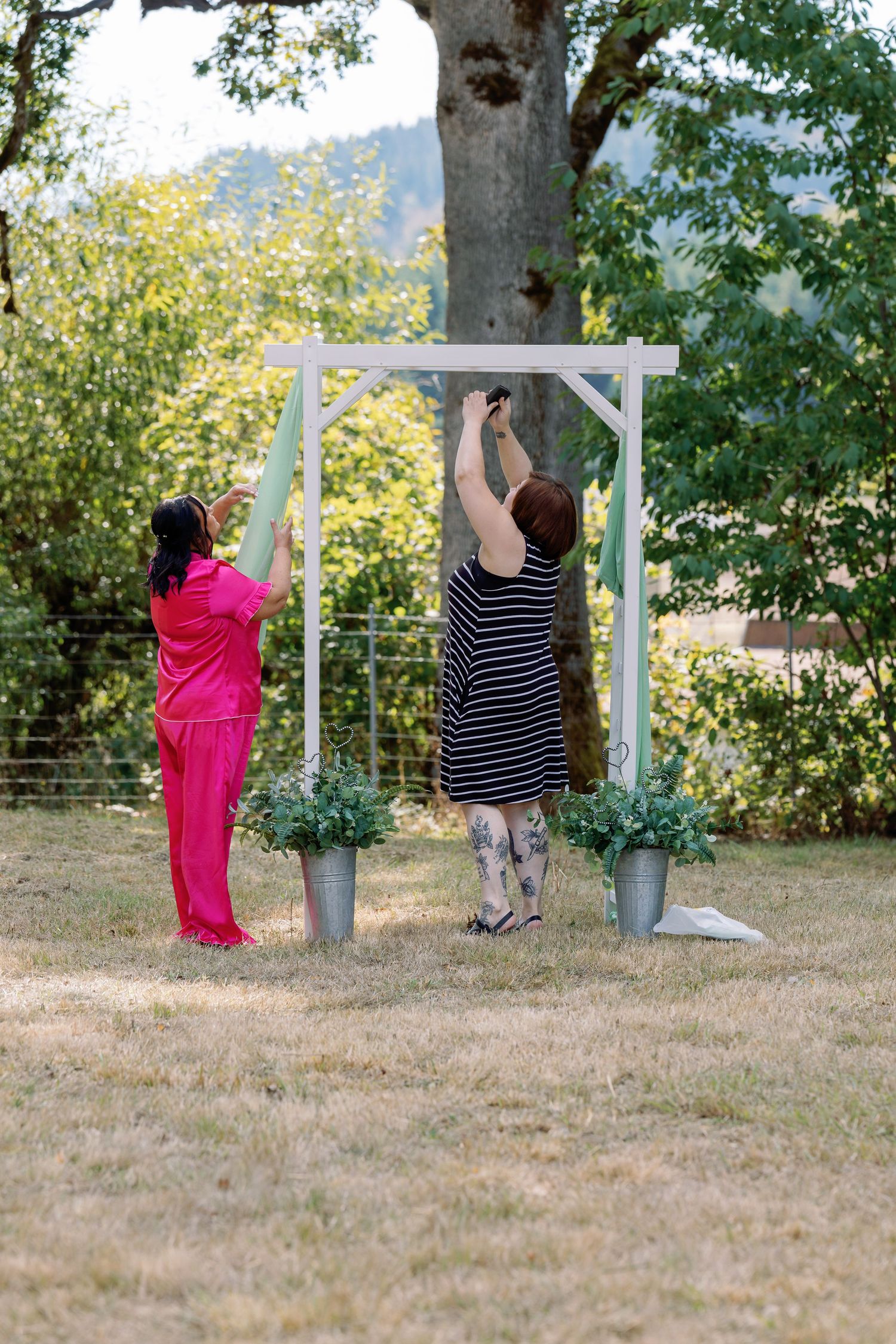 People decorating a white wooden arch in a garden setting with mountains visible in the background.