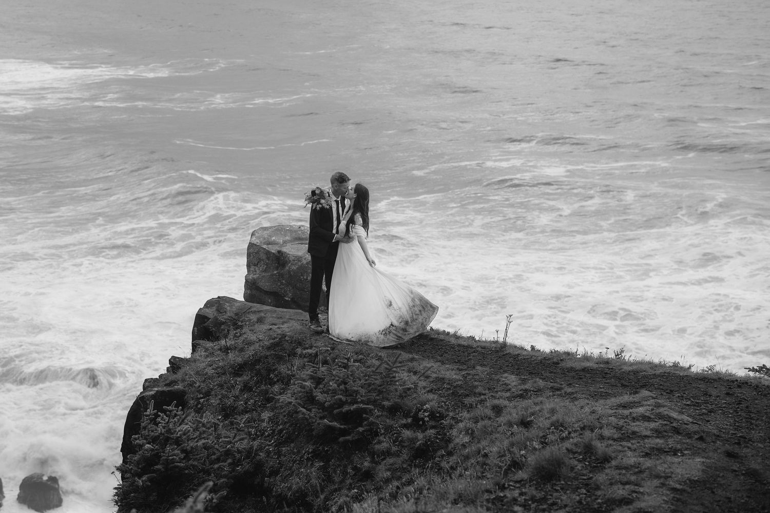 A couple stands together on a dramatic cliffside with crashing waves and ocean views below.