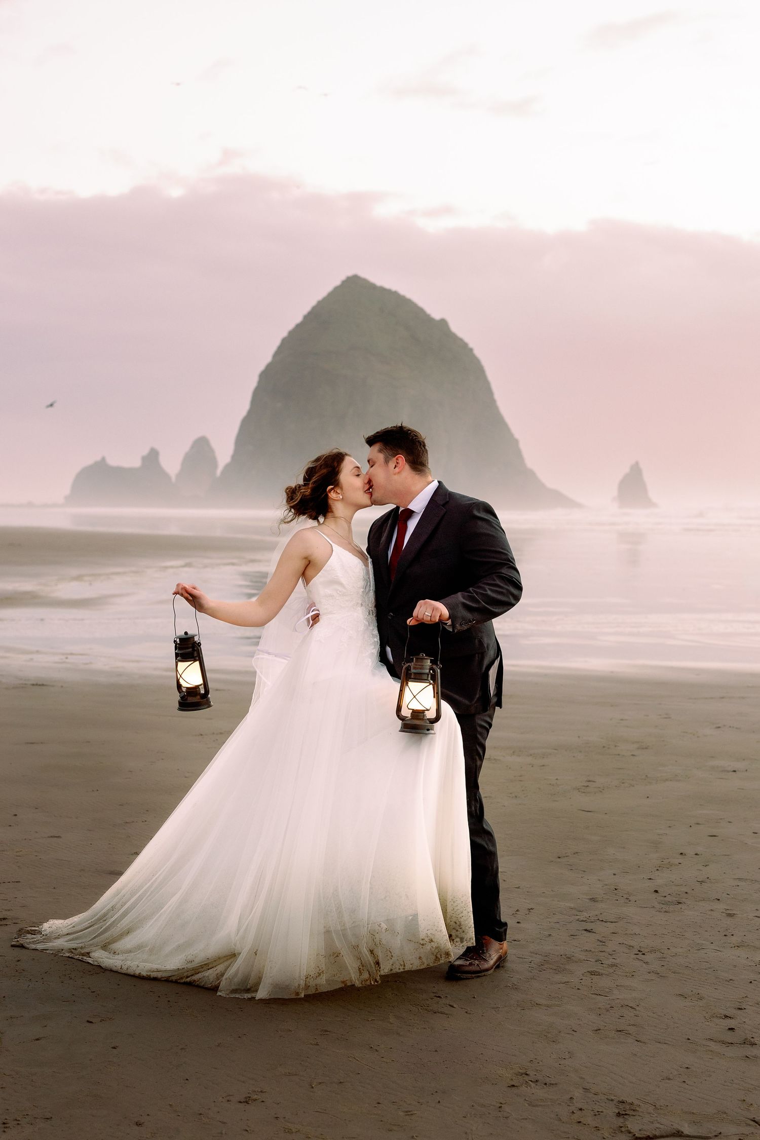 A romantic wedding couple dances on a misty beach with dramatic coastal cliffs in the background at sunset.