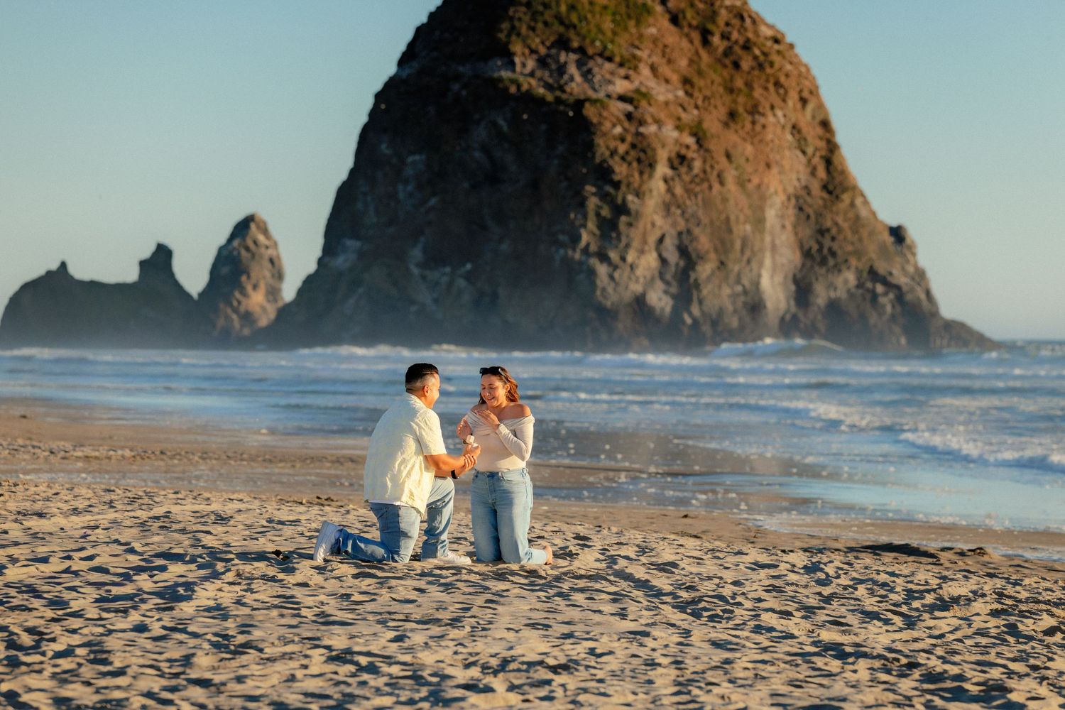 Couple sharing a romantic beach proposal moment at Cannon Beach with iconic Haystack Rock in the background.