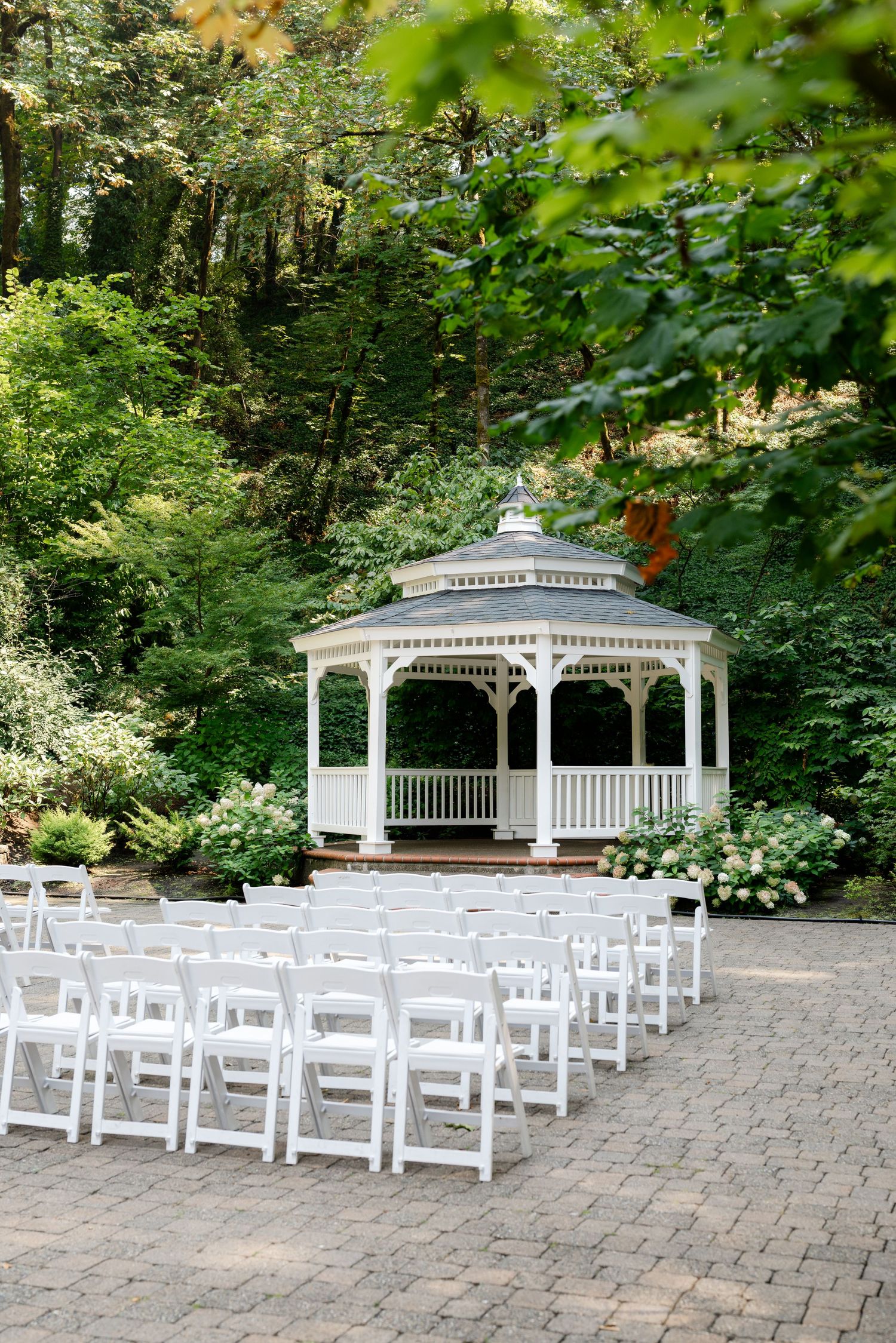 Elegant white gazebo surrounded by lush greenery with white folding chairs arranged for an outdoor wedding ceremony.