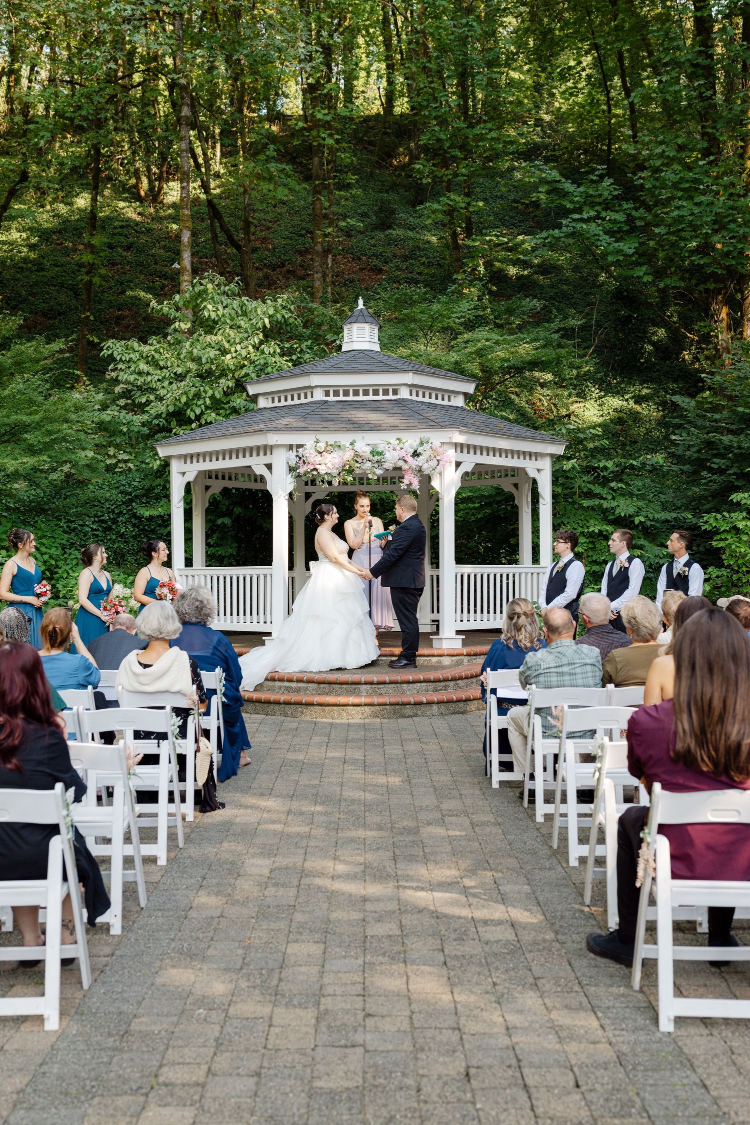 Outdoor wedding ceremony at a white gazebo surrounded by lush greenery with guests seated on white chairs.