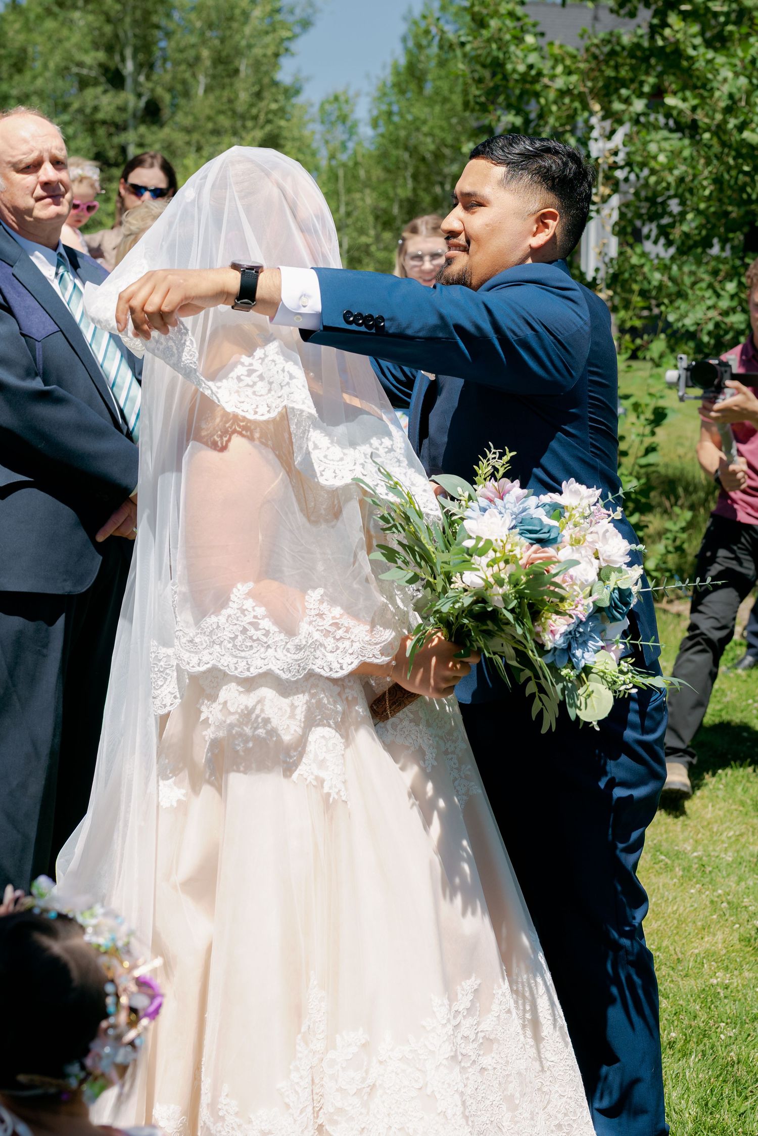 Groom in navy suit lifting bride's lace veil during outdoor wedding ceremony.