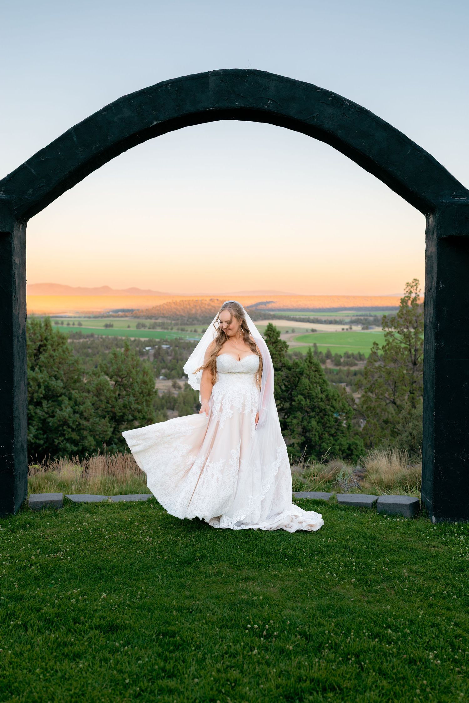 Elegant bride in a flowing white gown and veil stands under a stone arch at sunset with scenic valley views.