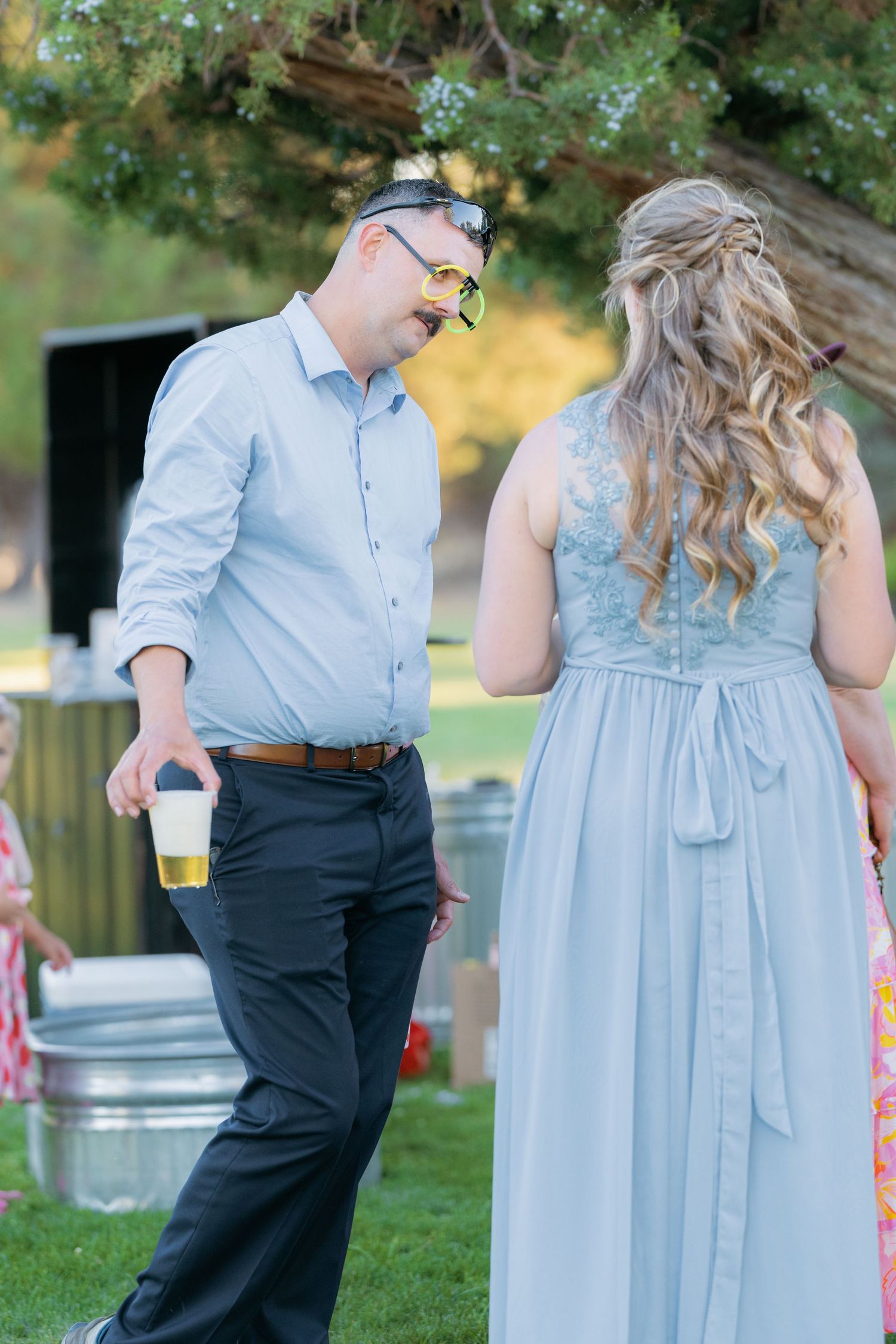 Man in light blue shirt holding a drink chats with woman in blue gown at outdoor wedding reception.