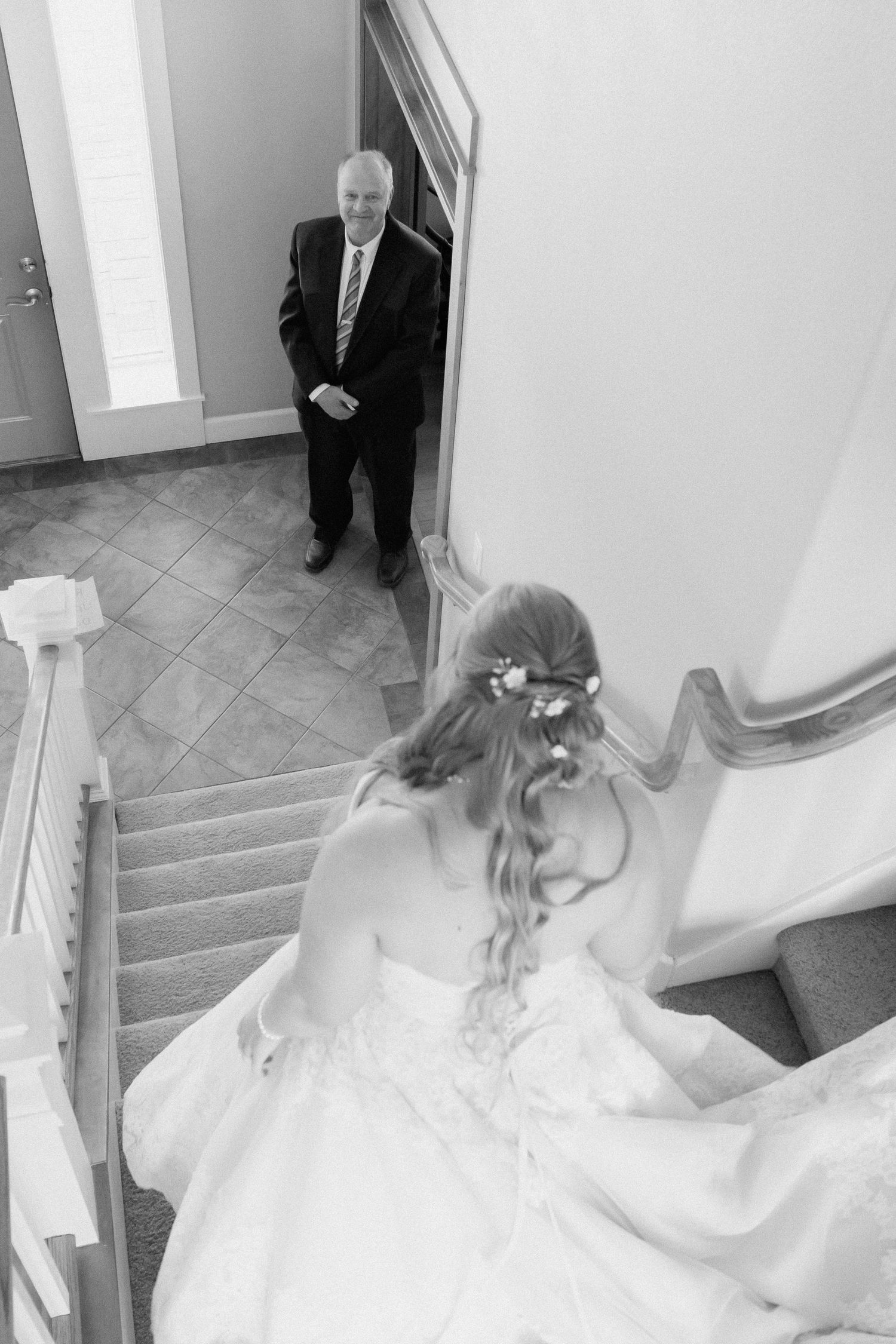 Black and white photo of a bride descending stairs in wedding gown while a man in suit watches from below.