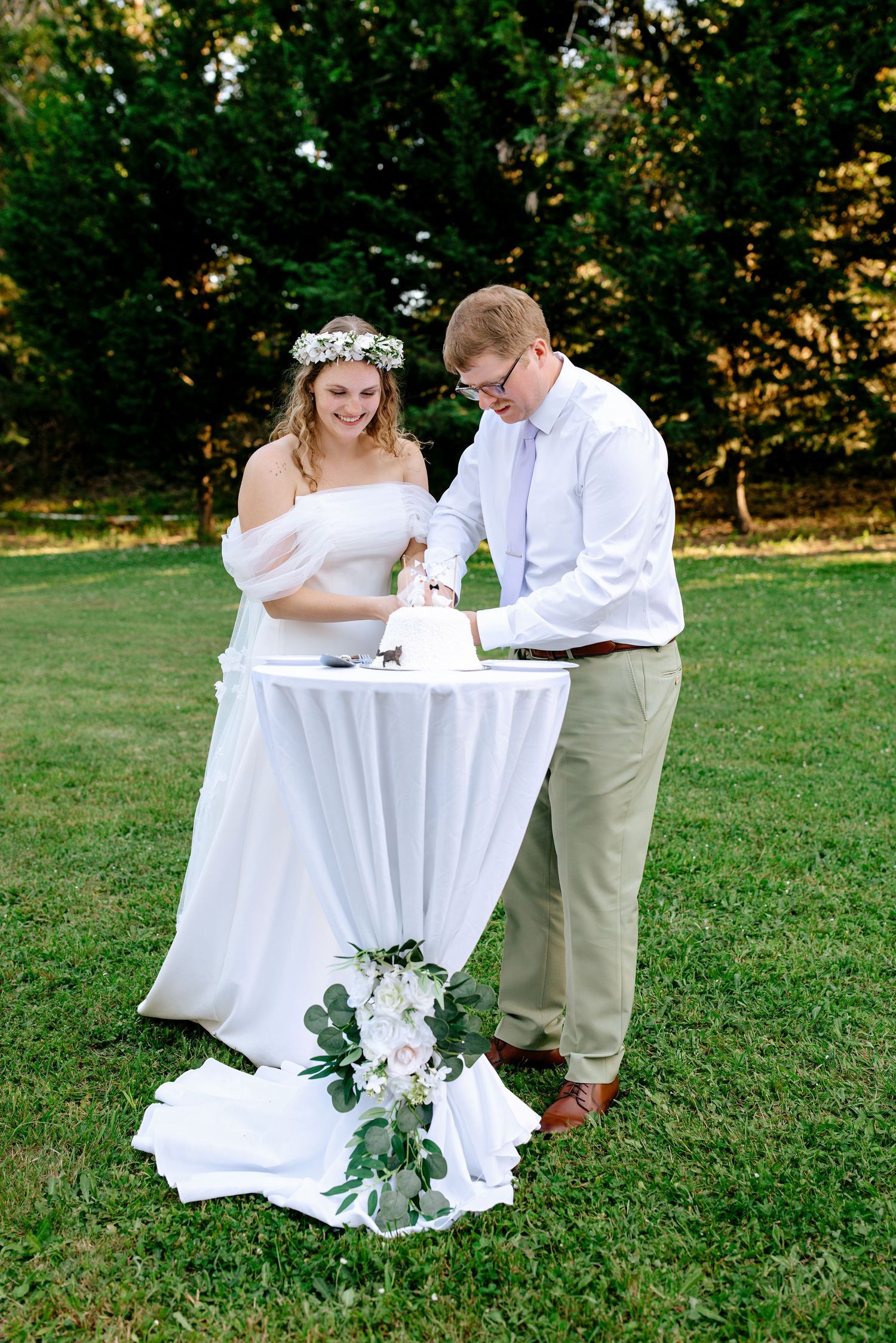 Bride in white gown and floral crown cuts wedding cake with groom in sage suit on a green lawn.