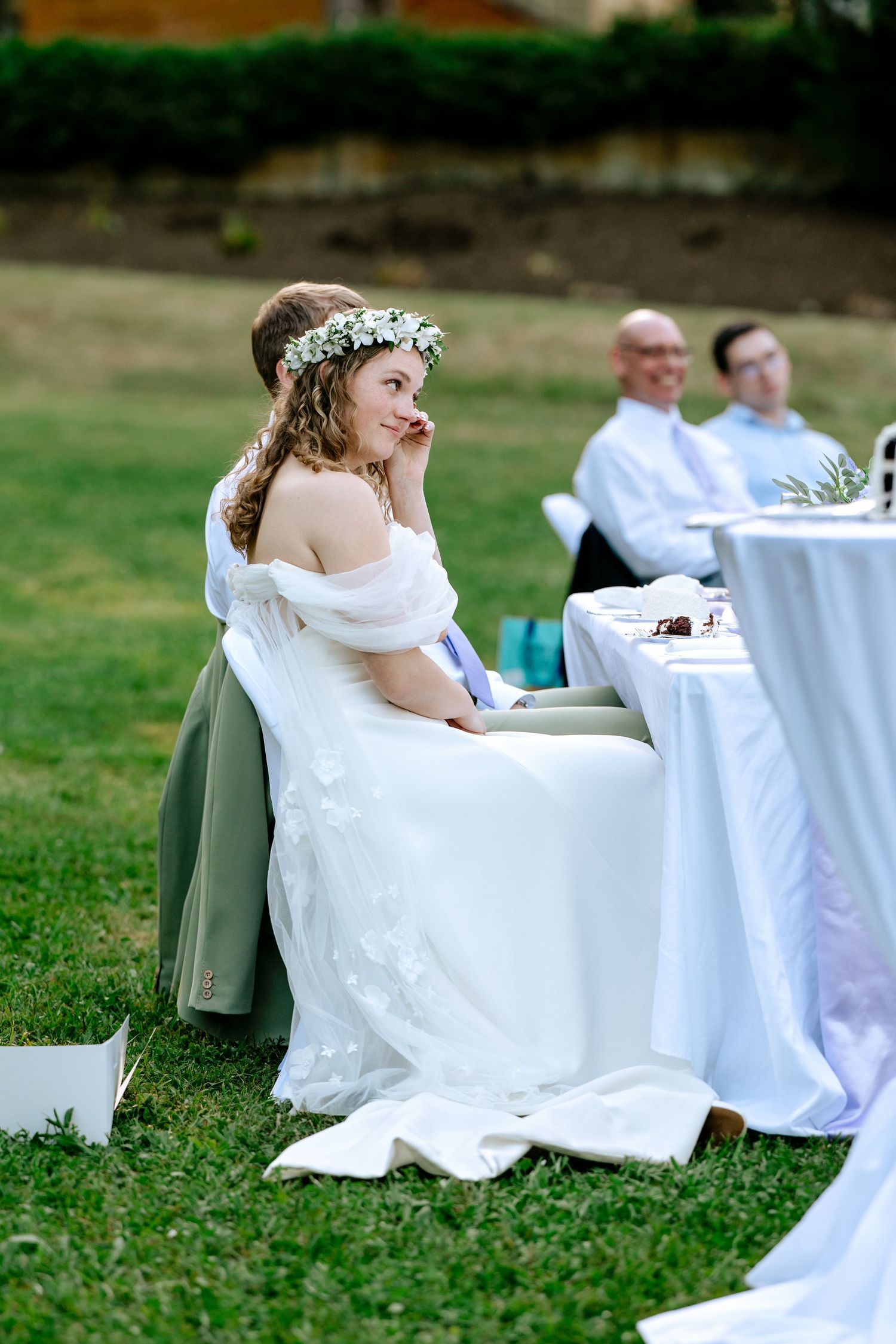 Bride in white gown and floral crown sitting at outdoor wedding reception table.