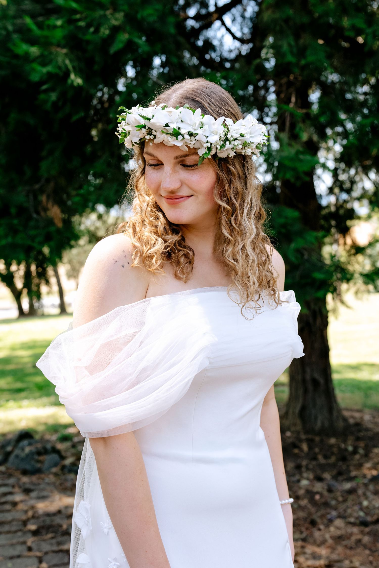 Smiling bride in white off-shoulder dress wearing a floral crown of white flowers in a park setting.
