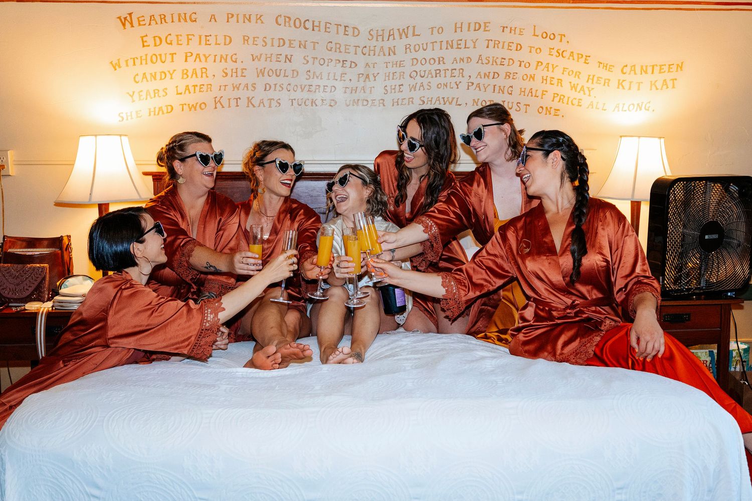 Bridesmaids in rust satin robes toasting drinks on a white bed, wearing sunglasses and smiling together.