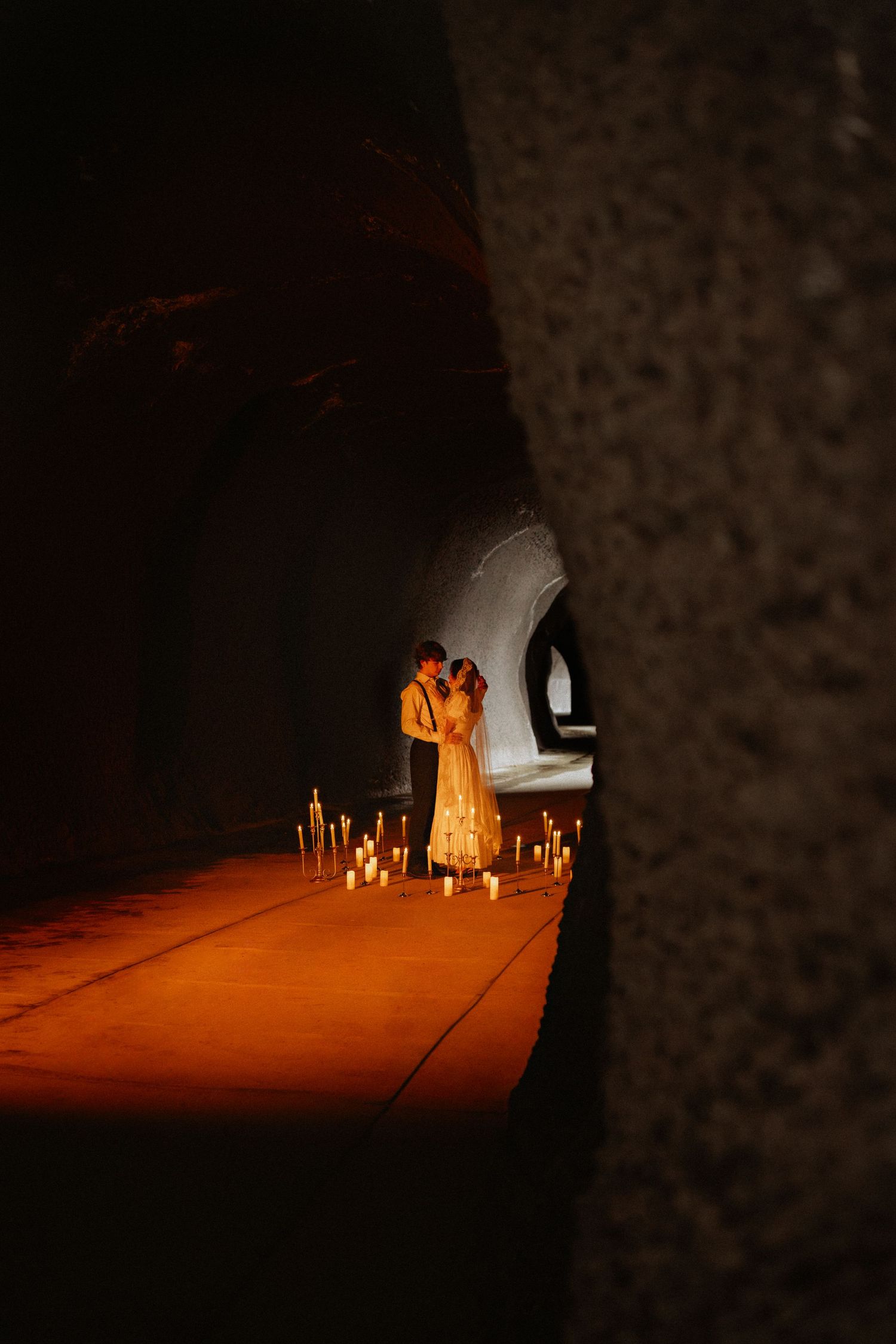 A couple embracing in a dimly lit stone tunnel surrounded by flickering candles.