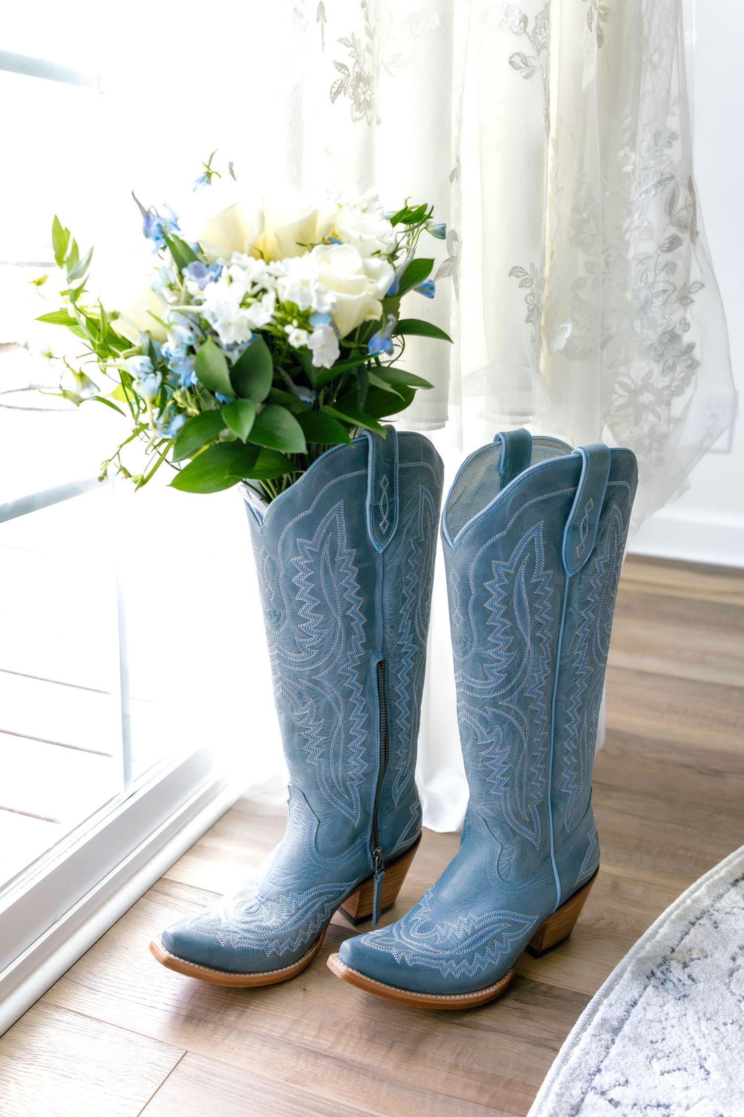 Light blue cowboy boots with embroidered details sit next to a white floral bouquet near a lace curtain.
