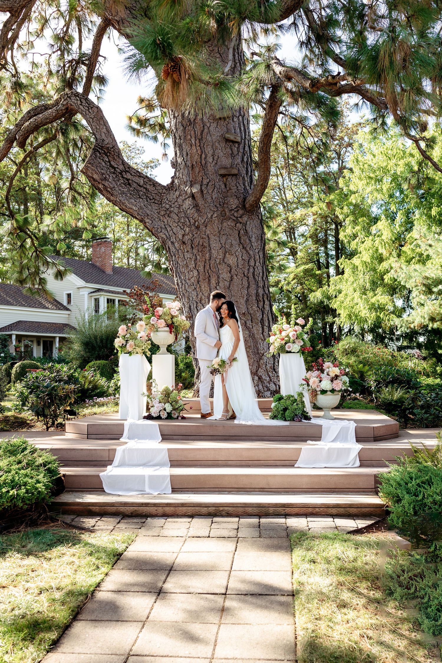 A couple dancing and twirling in white attire on a garden patio with wooden steps and large pine trees.