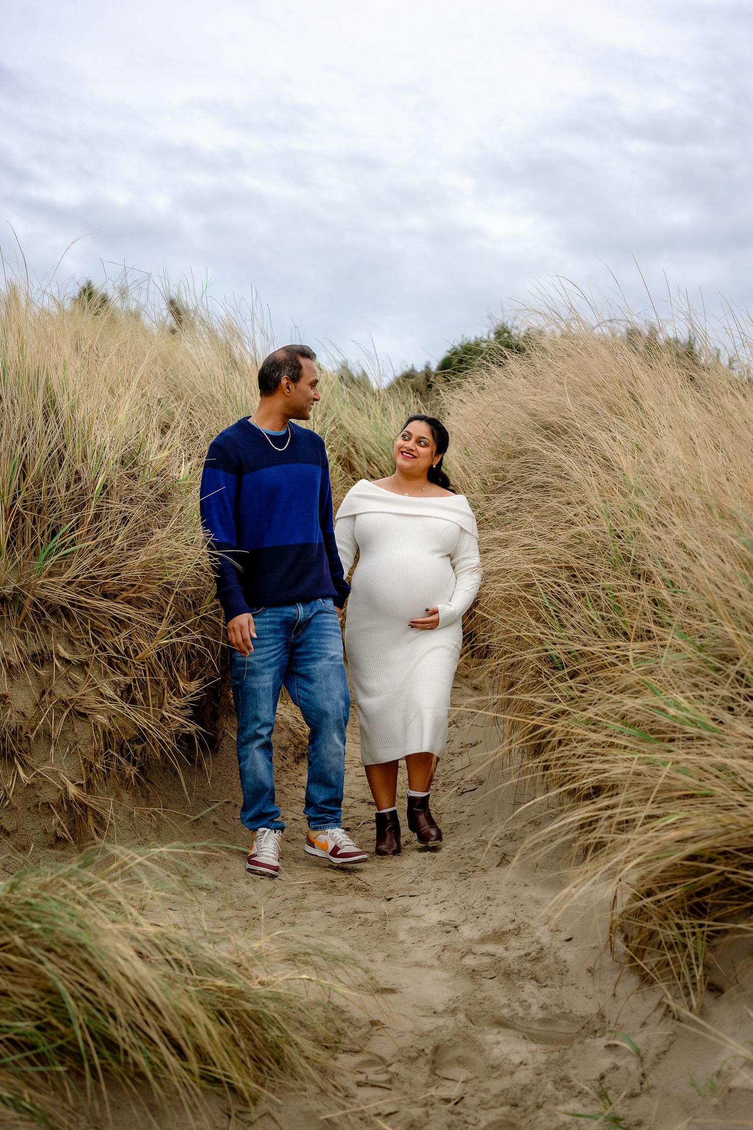 A couple shares an intimate moment among tall beach grass on sandy dunes under an overcast sky.