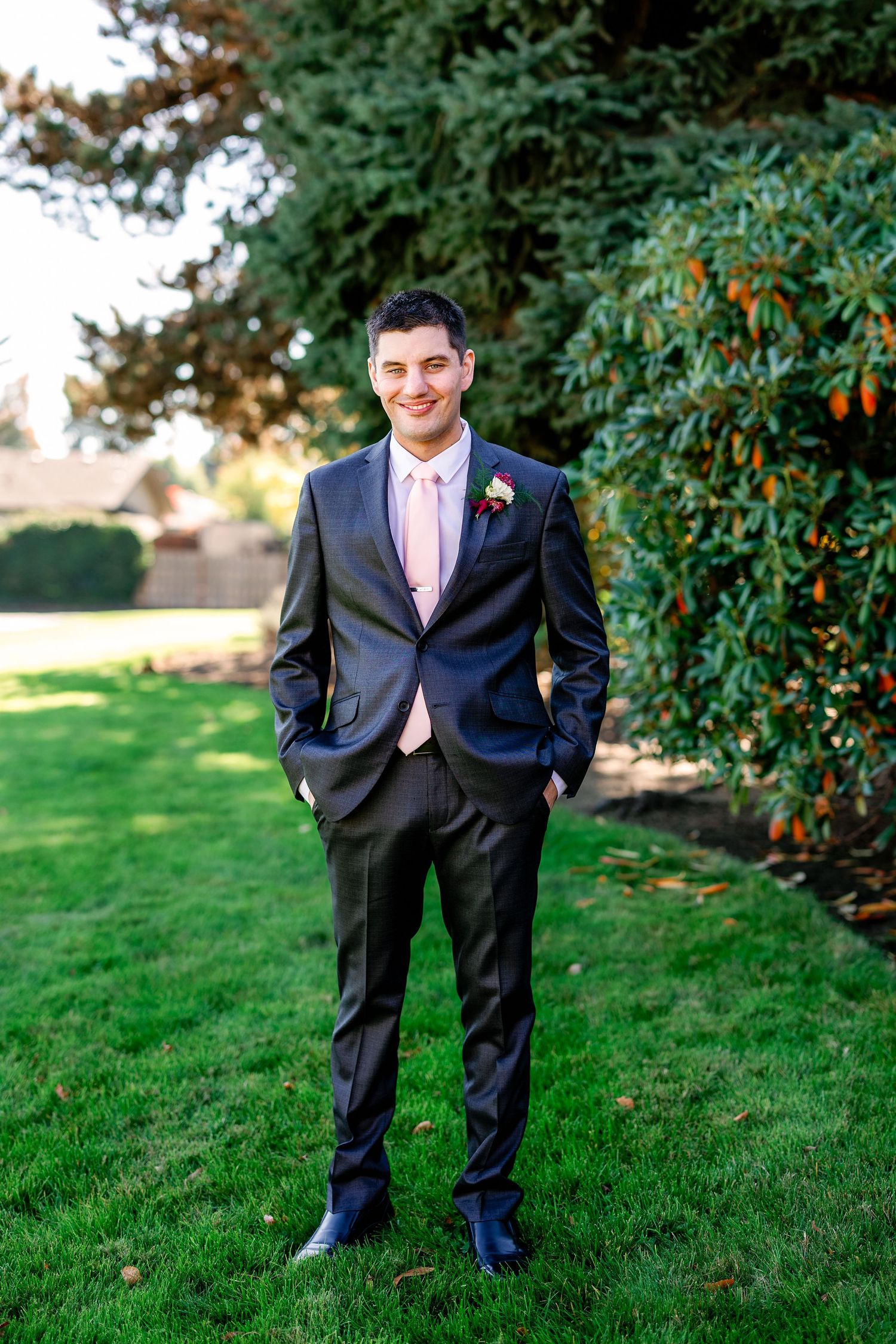 Groomsman in black suit with pink tie poses on grass near garden foliage.