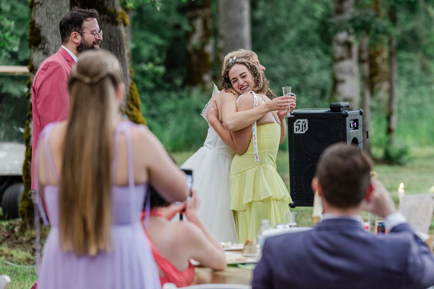 People gather for an outdoor celebration in a wooded setting with attendees in formal attire enjoying the festivities.