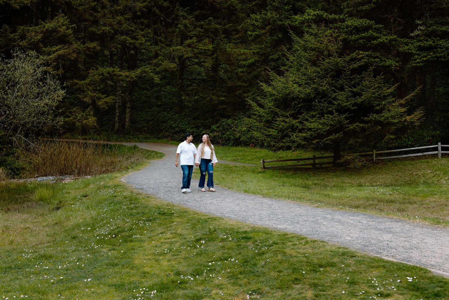 Couple walking hand in hand on a scenic gravel path surrounded by lush green grass and dense forest trees.