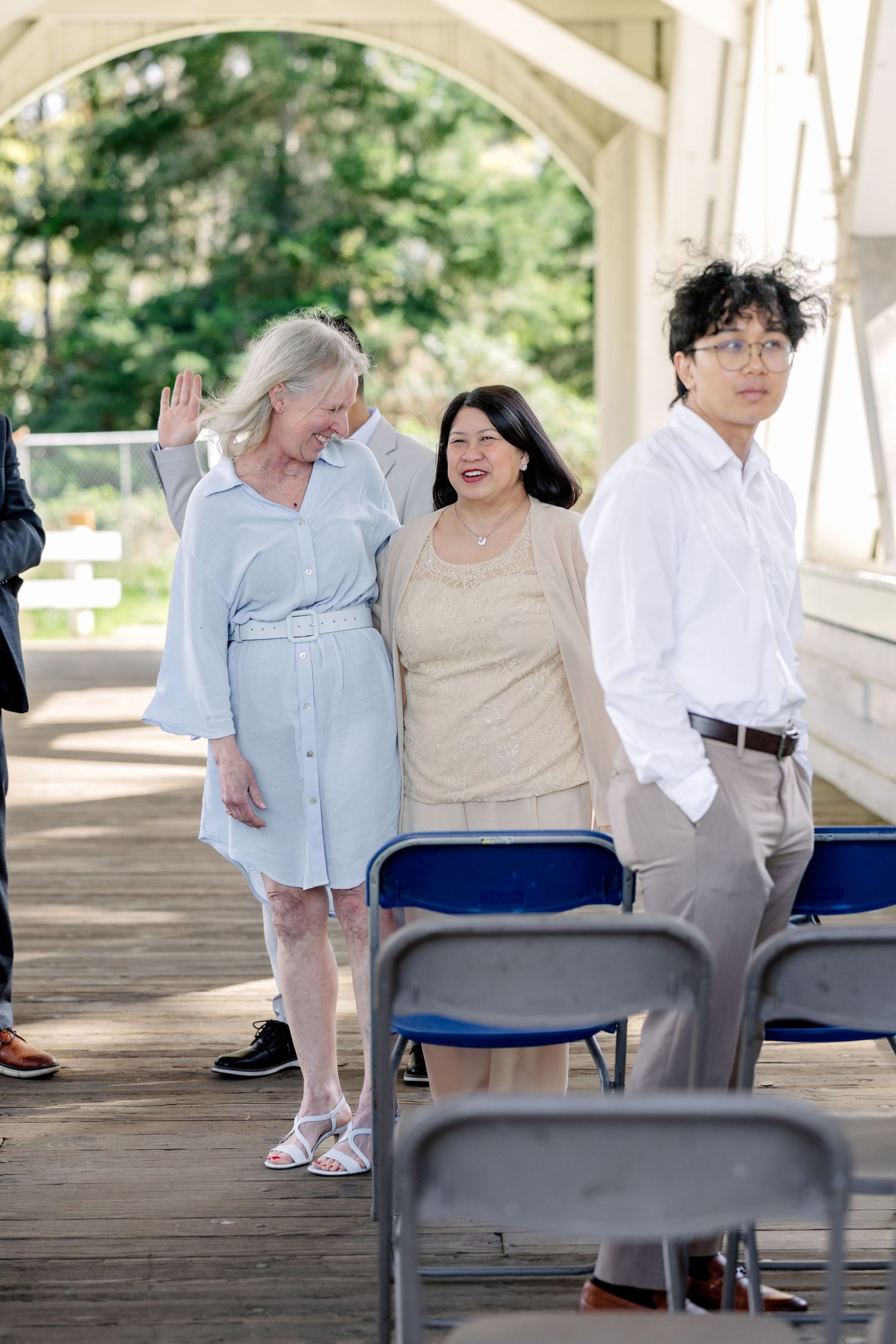 Wedding guests smiling and waving during an outdoor ceremony on a wooden bridge.