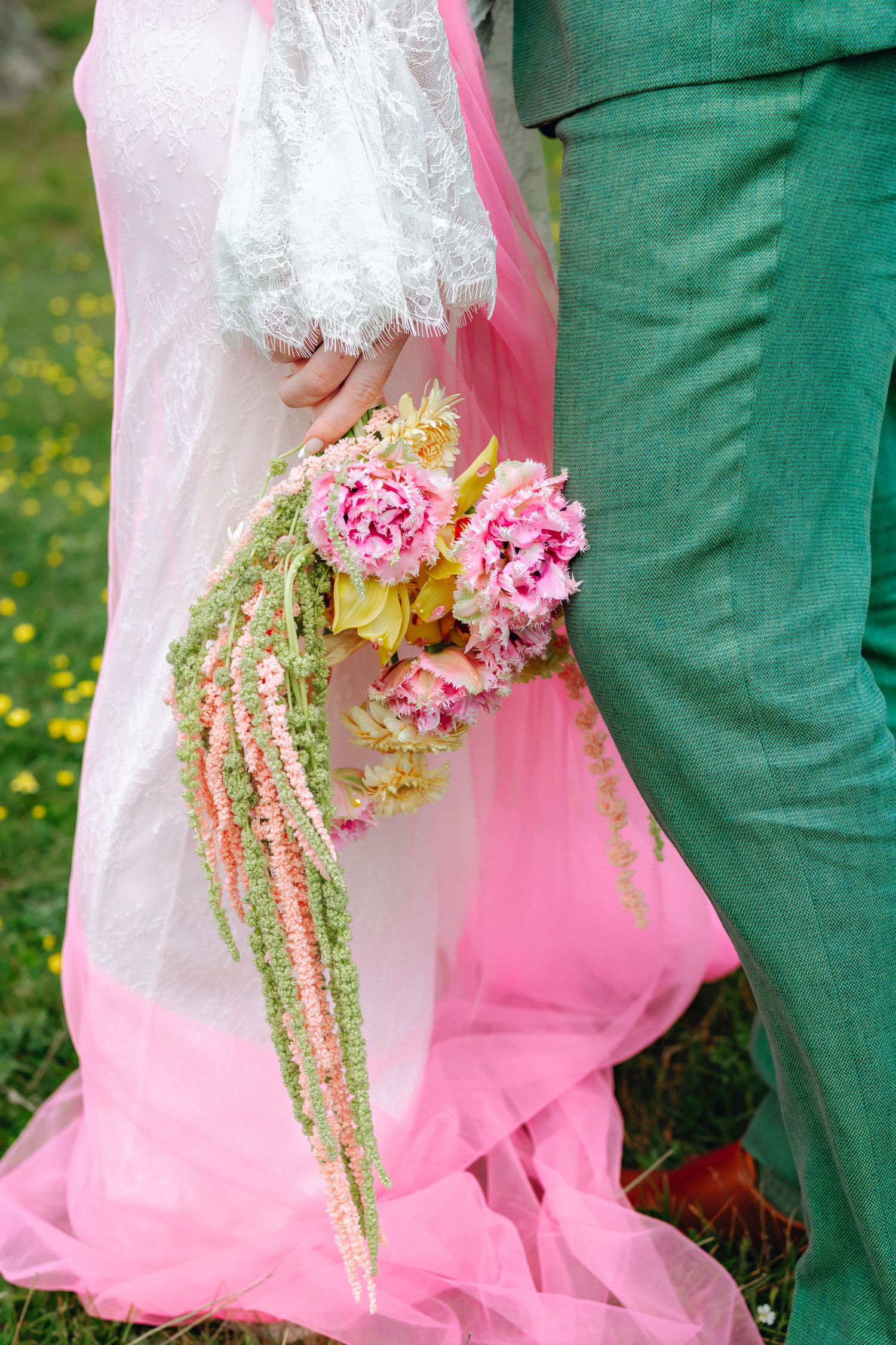 Bride in white and pink dress holds colorful bouquet of pink peonies beside groom in green suit.