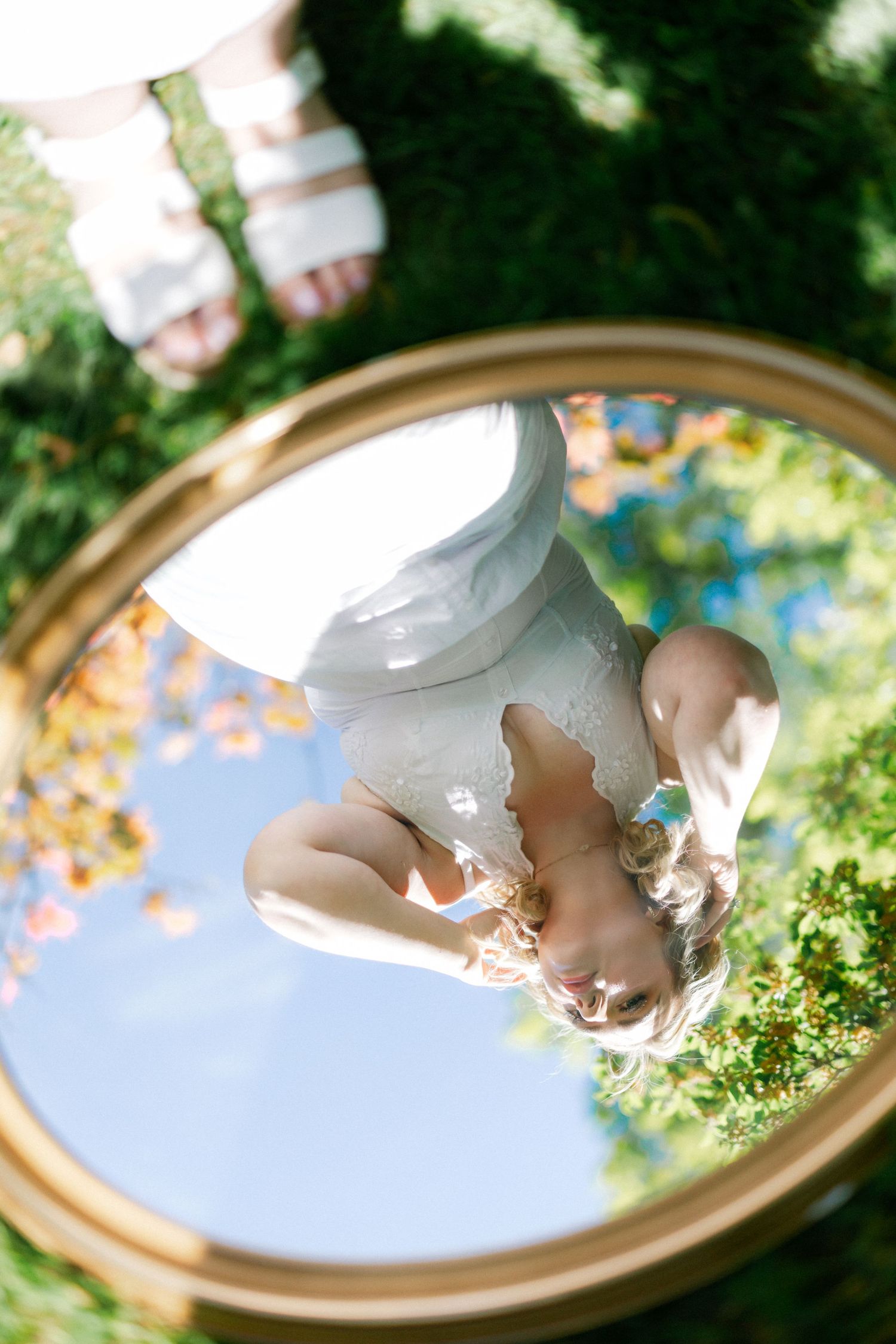 Woman in white dress reflected upside down in a round gold mirror outdoors among green grass and trees.