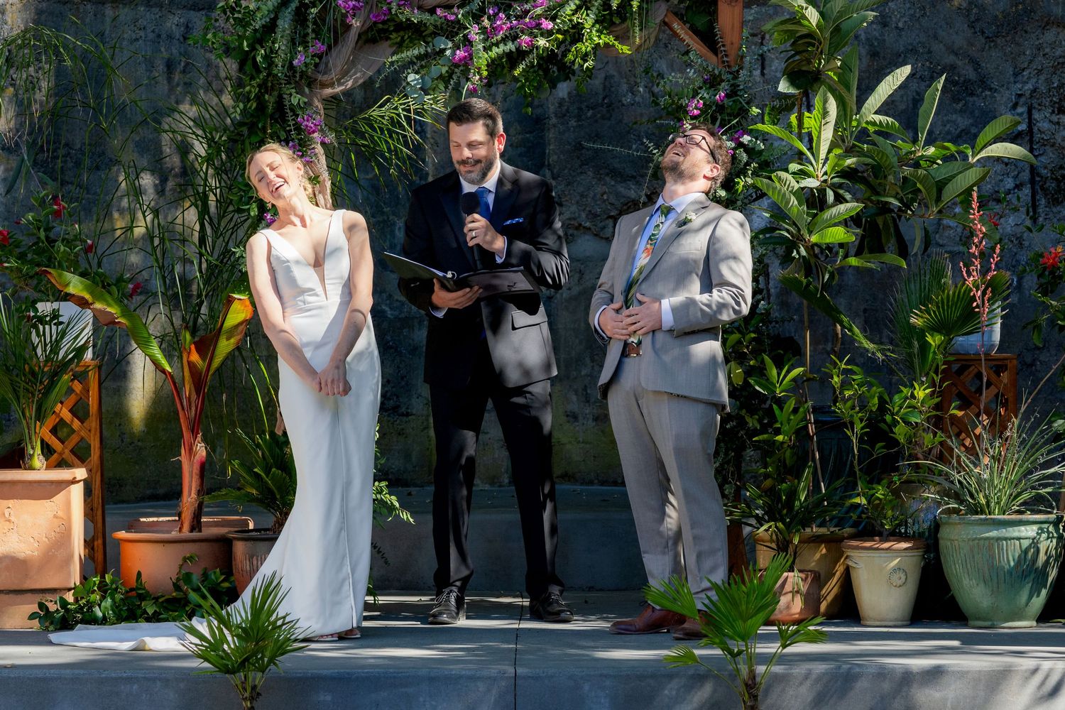 Couple exchanges vows at outdoor wedding ceremony with officiant, surrounded by lush tropical plants.