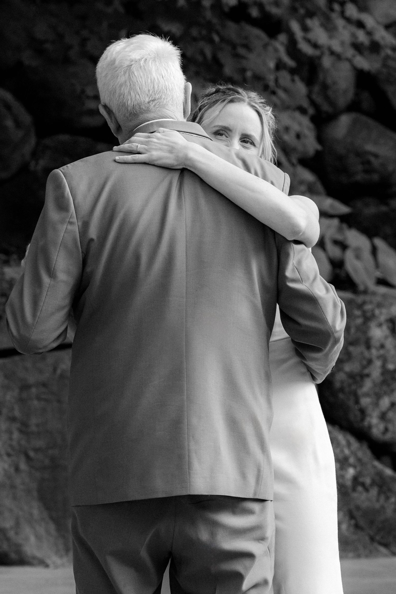 Black and white photo of bride embracing older man in suit at outdoor wedding ceremony.