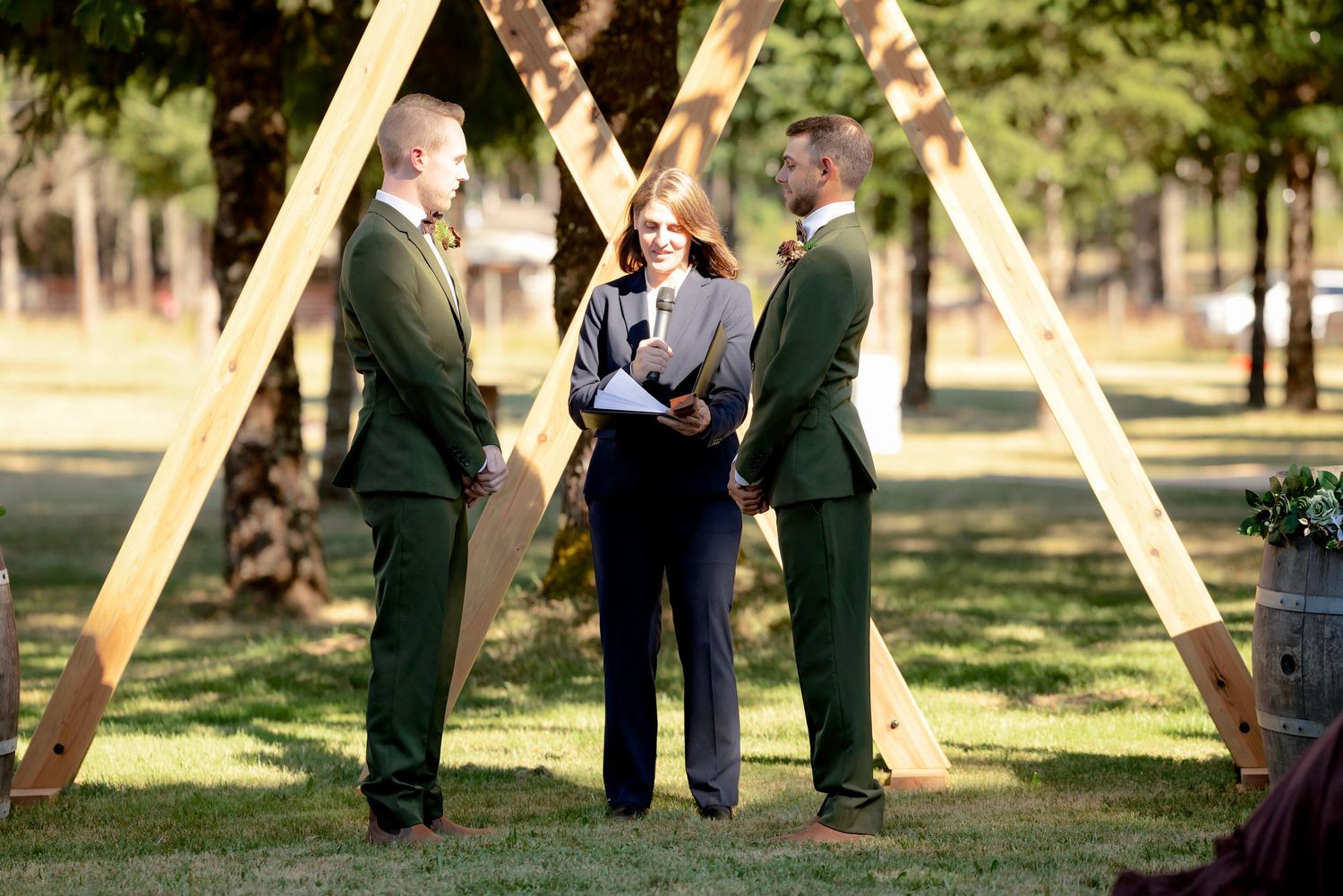 Two grooms in dark green suits exchanging vows under wooden geometric arch with officiant at outdoor wedding ceremony.