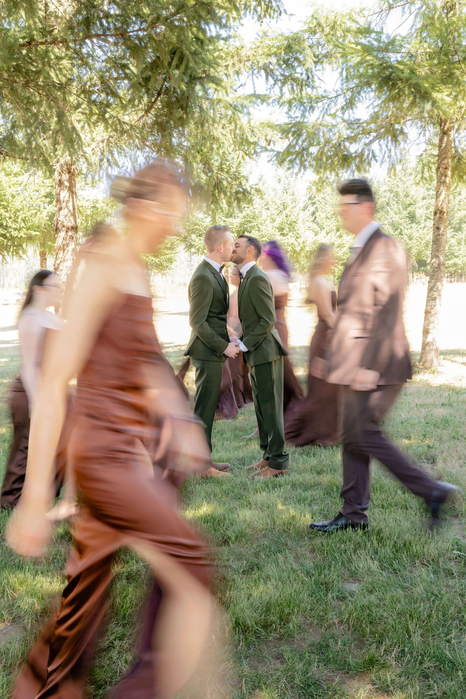 Artistic motion blur photo of two grooms kissing in olive suits surrounded by moving wedding party guests under pine trees.