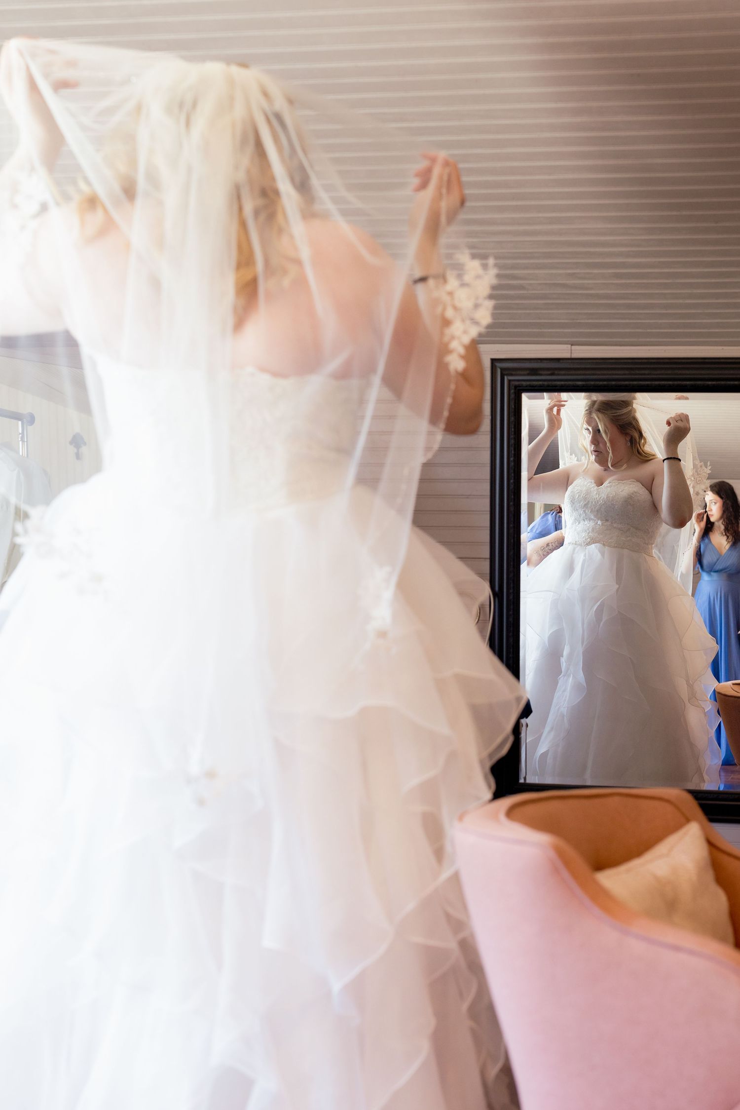 Bride in white ruffled gown adjusting veil before mirror with bridesmaid reflection visible.