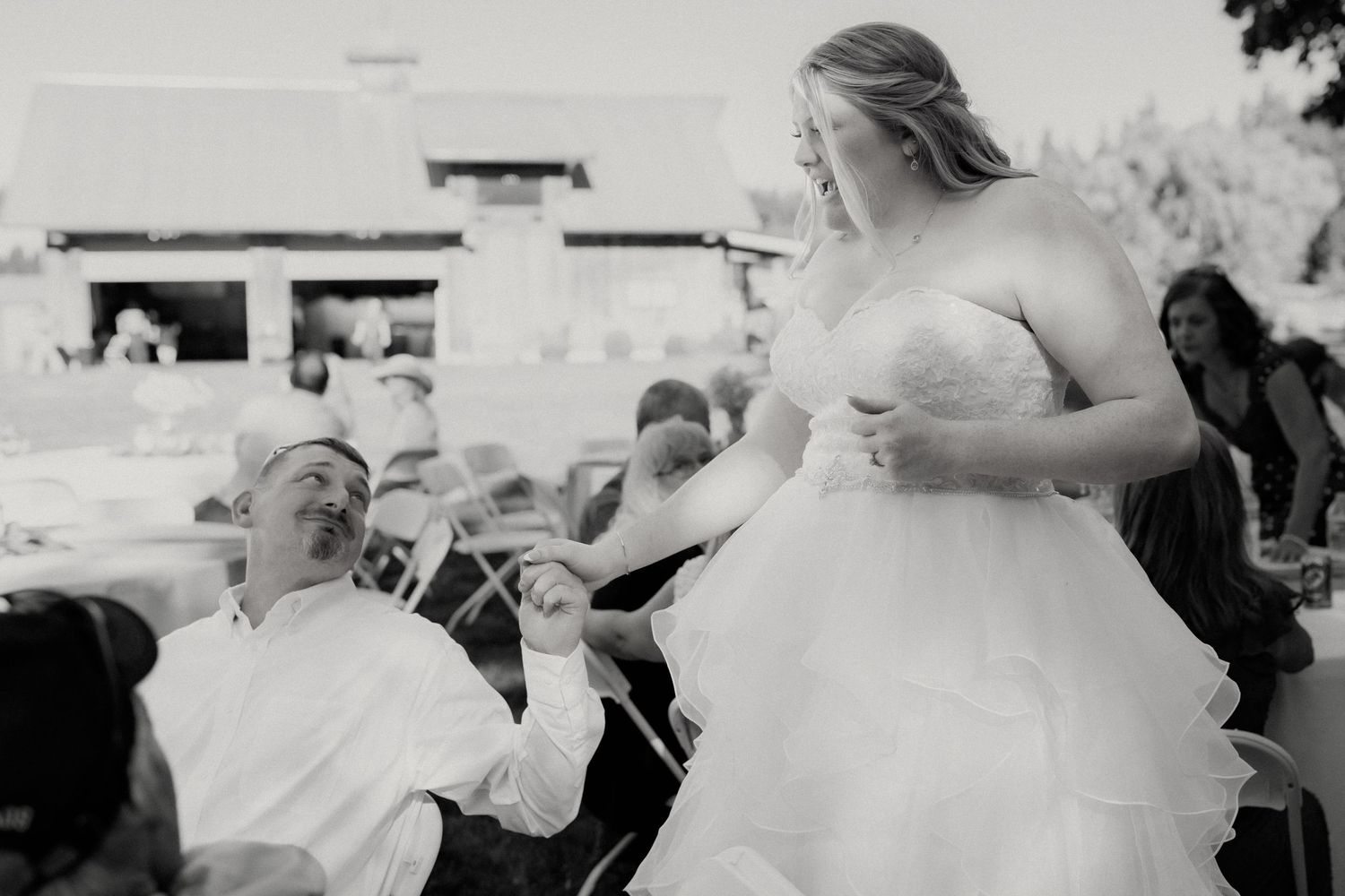 Black and white photo of bride in strapless gown interacting with seated guest at outdoor wedding.