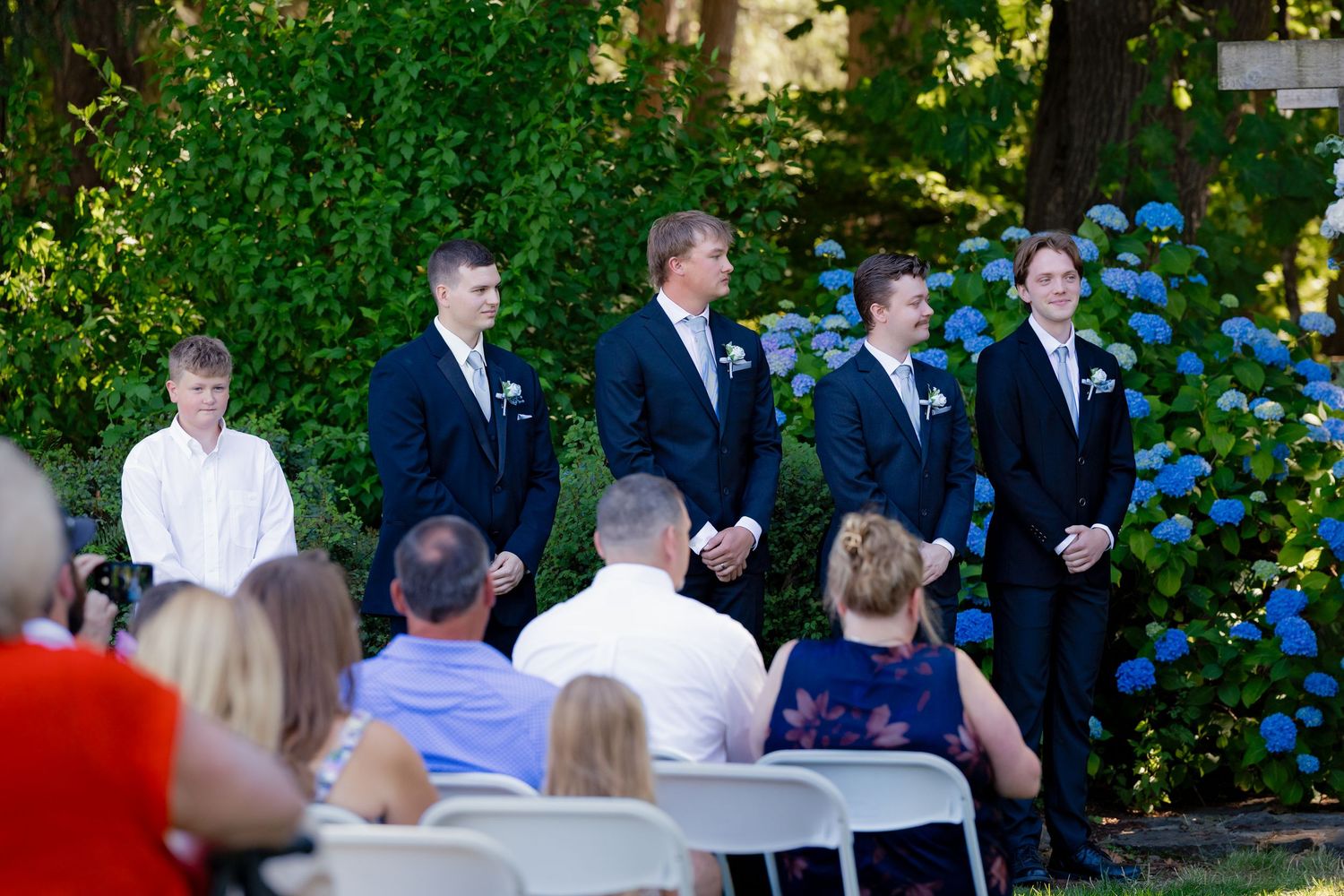 Groomsmen in navy suits standing at an outdoor wedding ceremony with seated guests and hydrangeas in background.