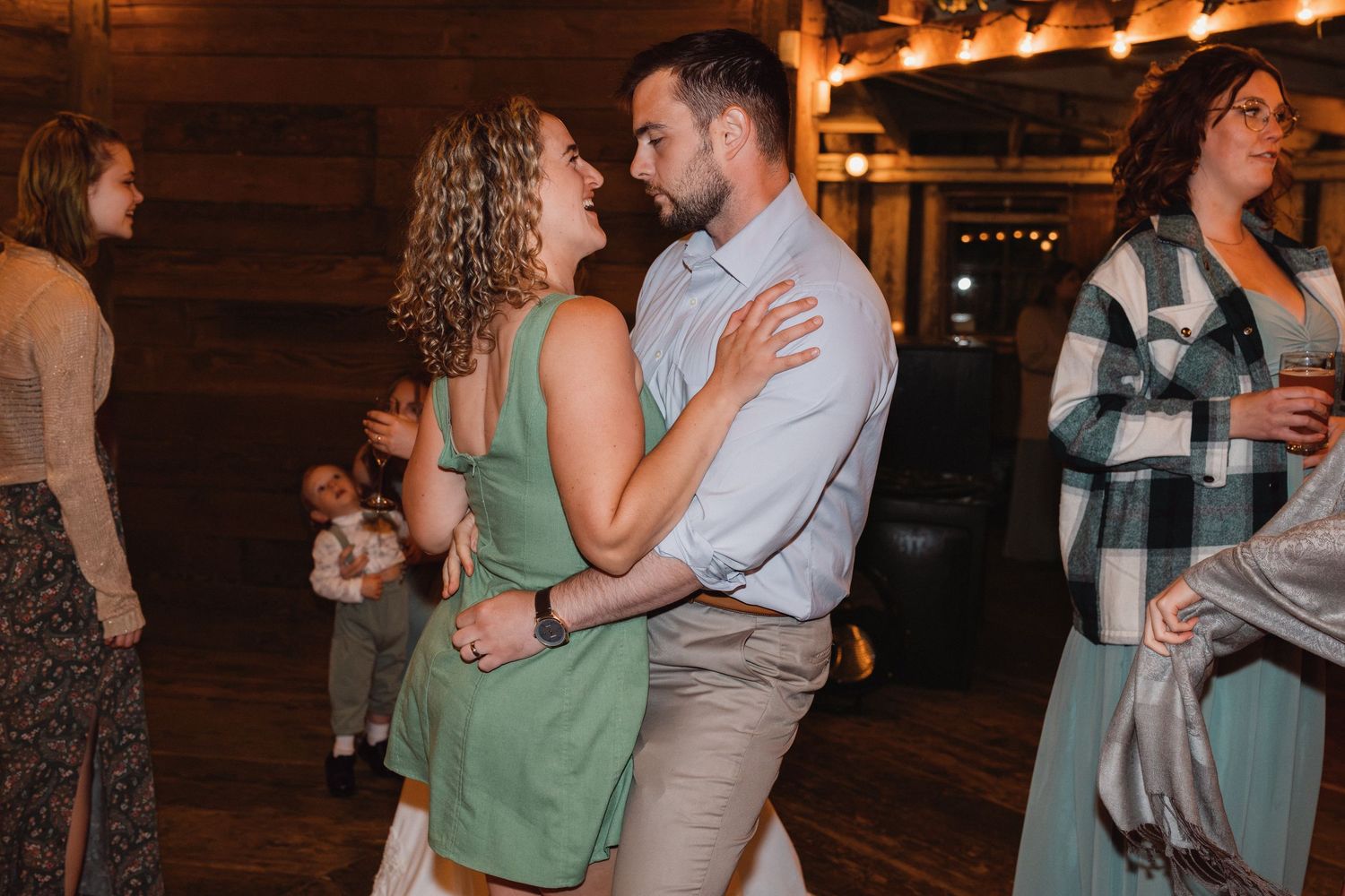 Couple slow dancing together at wedding reception in rustic barn venue with other guests nearby.