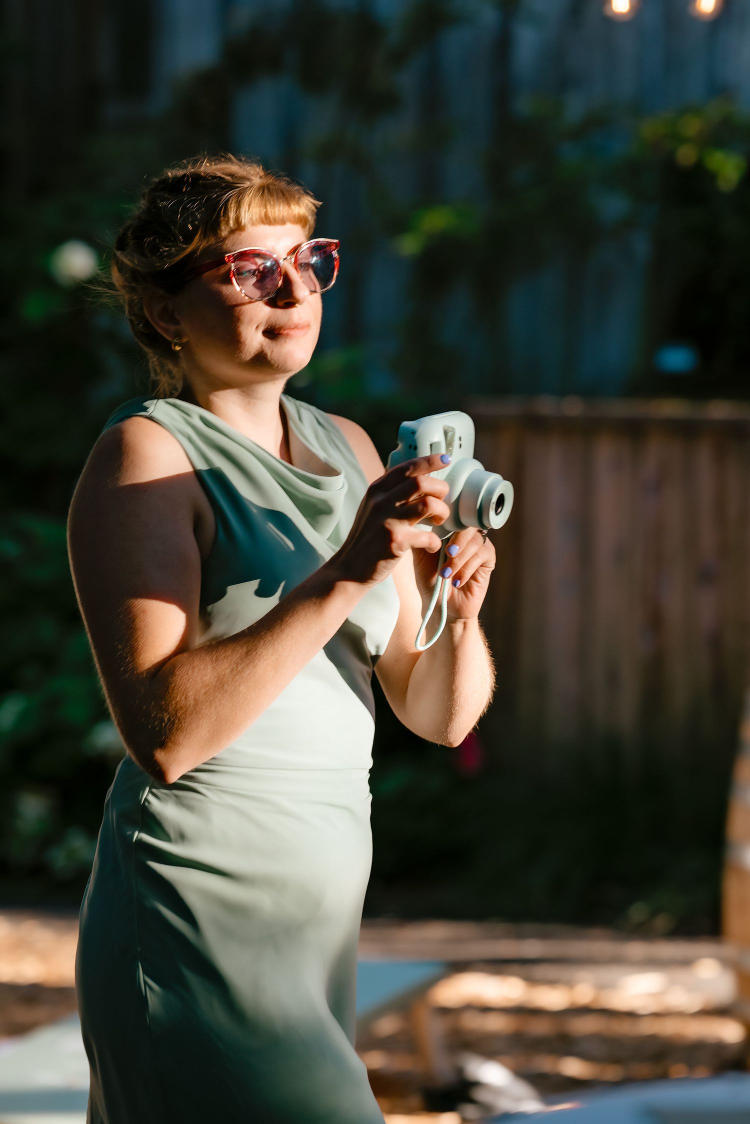 Stylish pregnant woman in sage green dress wearing pink sunglasses holds instant camera at outdoor evening event.