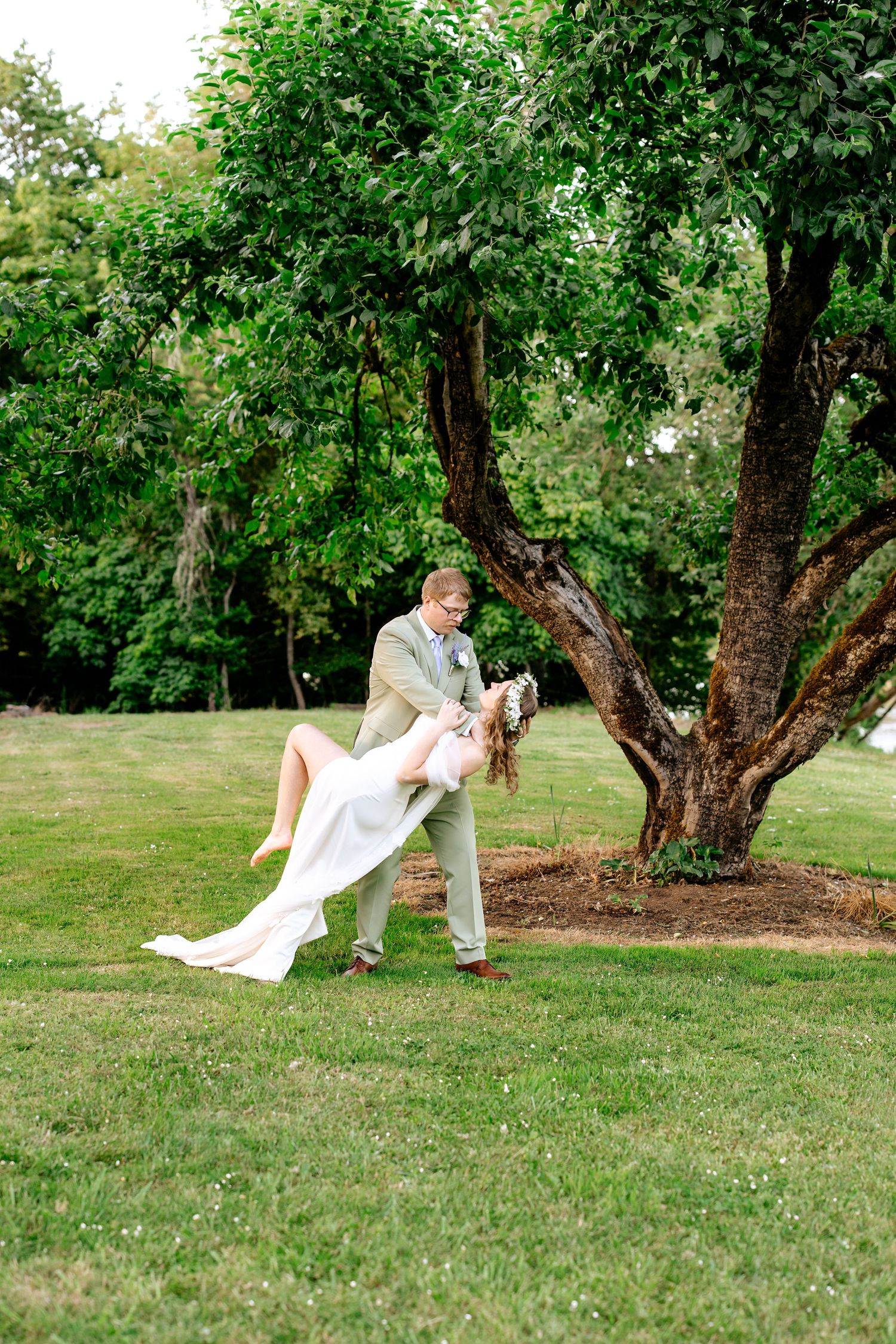 Groom in tan suit dips bride in white gown near a large tree on green lawn.