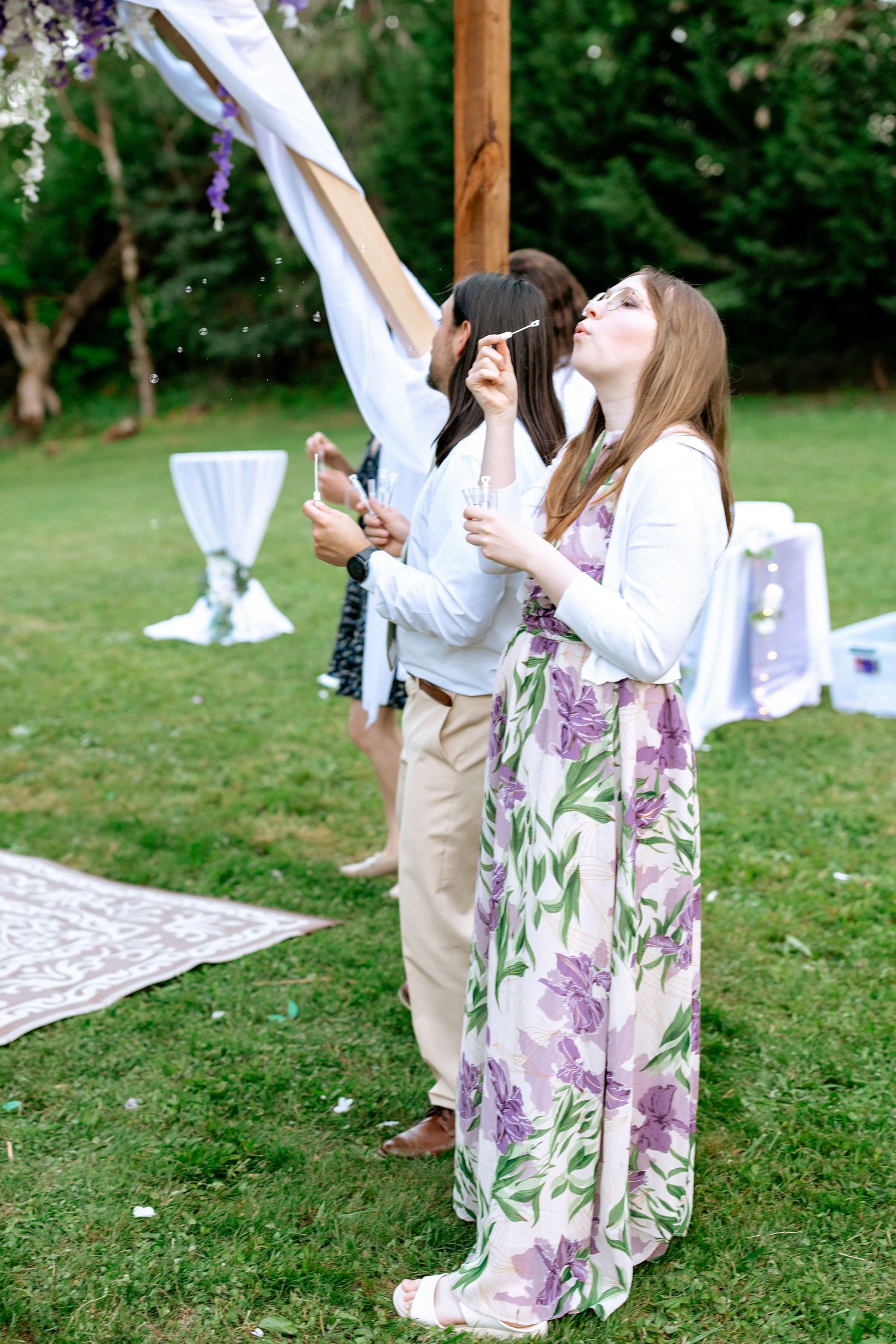 Two women in floral dresses blow bubbles near a decorated wooden arch at an outdoor garden wedding.