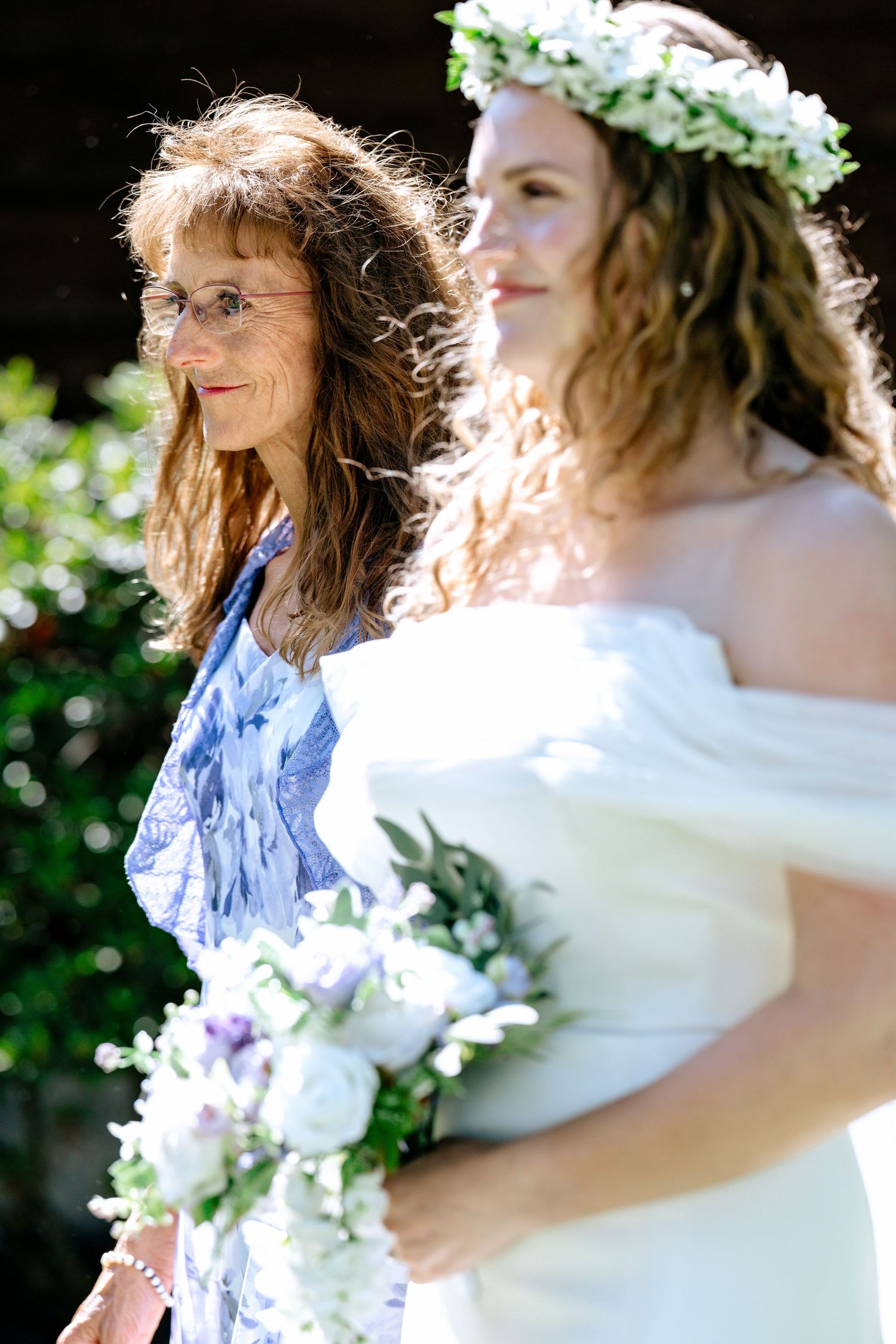 Bride in white off-shoulder dress with floral crown holds bouquet beside older woman in blue floral dress.
