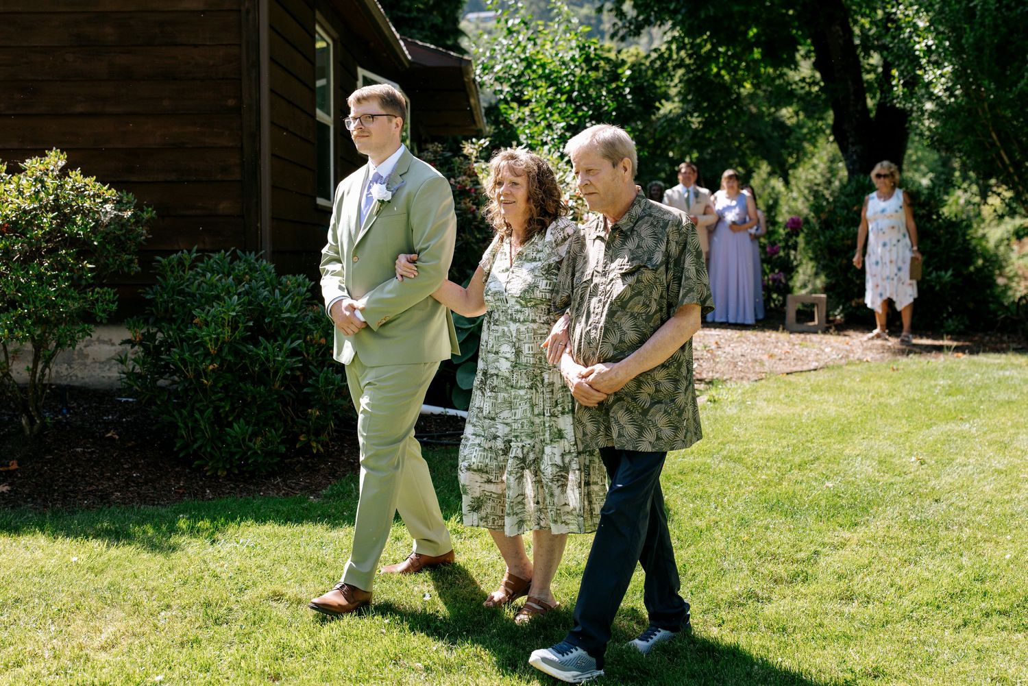 Groom in sage suit escorting two guests across green lawn at outdoor wedding venue.