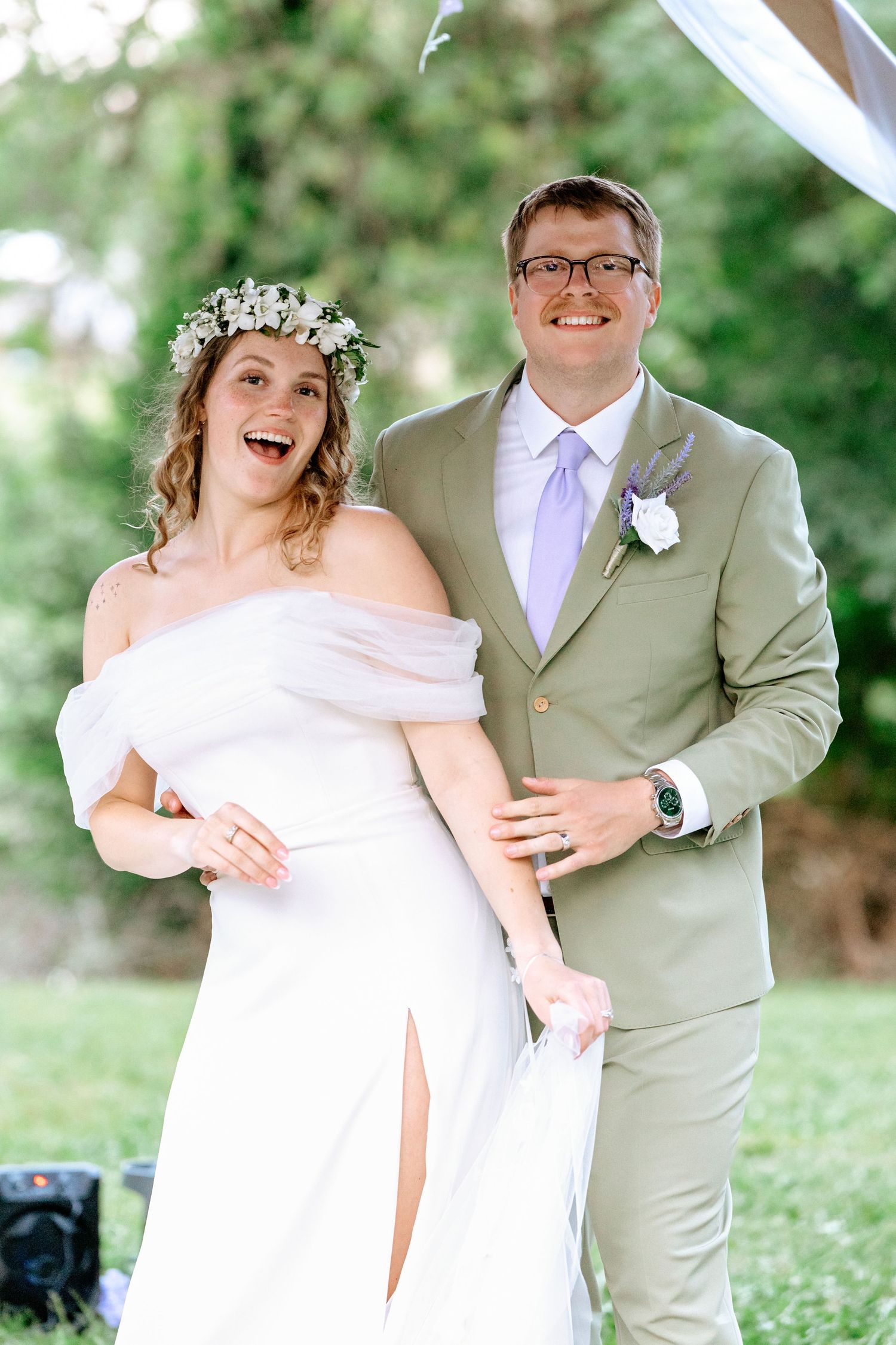 Joyful newlyweds laugh together after ceremony, bride in white gown with floral crown, groom in sage suit.